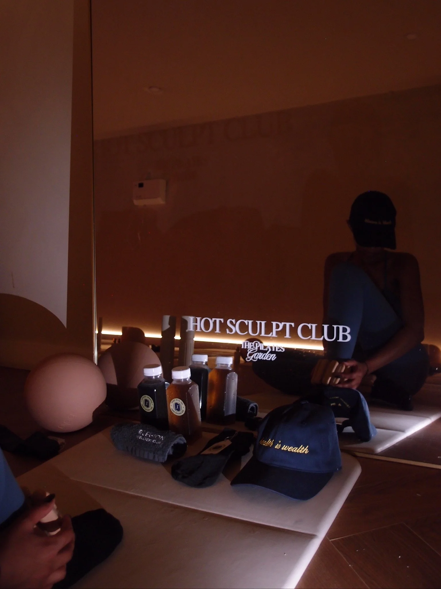 A mirror with bottles, hats, and other items displayed in front of it. Reflection shows a person taking the photo and a woman sitting on the floor. Text on the mirror reads 'HOT SCULPT CLUB' and 'THE PLATES Garden'.
