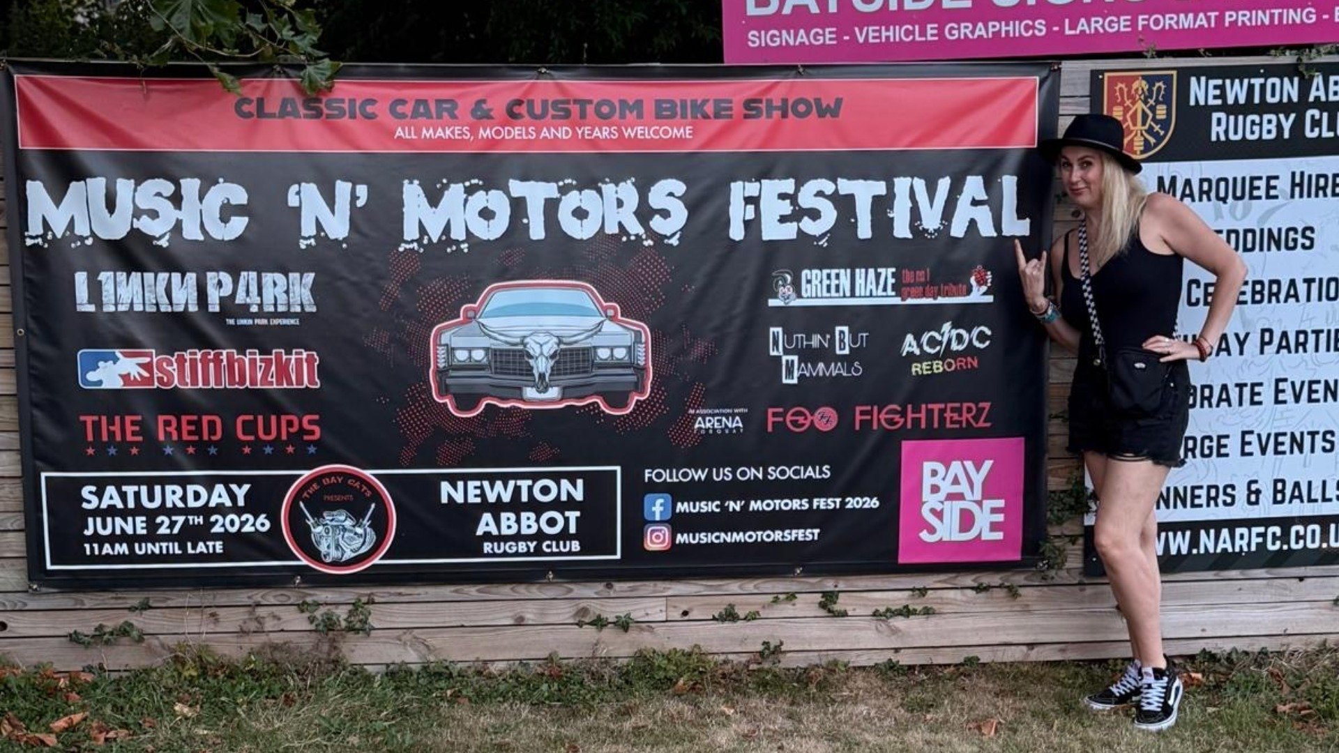 Julia beside the Music 'N' Motors Festival banner she designed, displayed at Newton Abbot Rugby Club ahead of the June 2026 event.