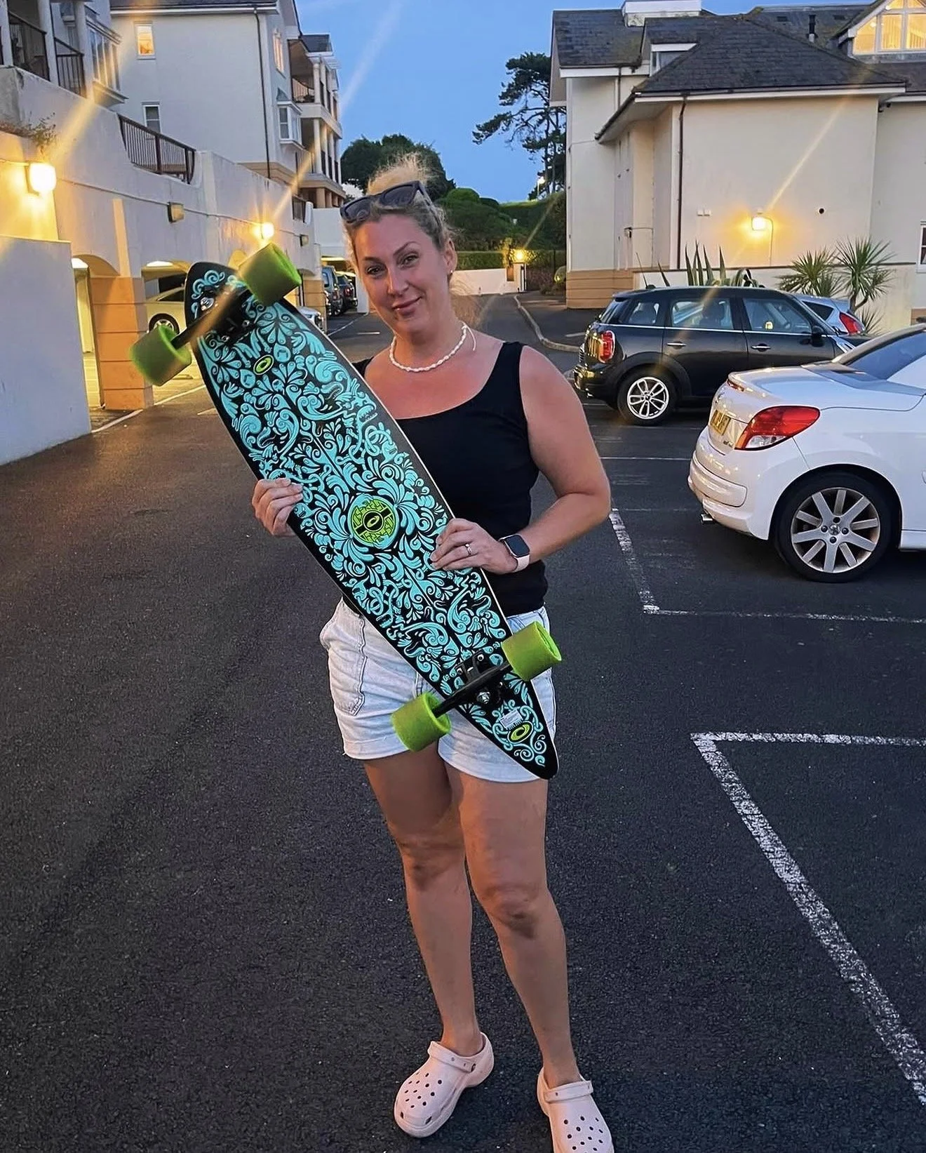 Julia holding an Osprey longboard in a car park at dusk, part of the 'outside the studio' section of the About page.