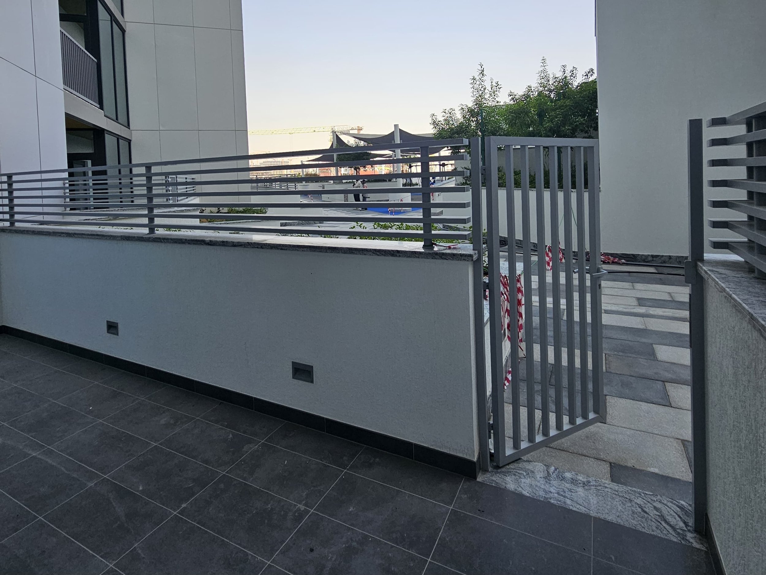 View of a modern balcony or terrace area with metal railing, gray tiles, and a gate leading to an outdoor space with construction barrier tape and a paved pathway.