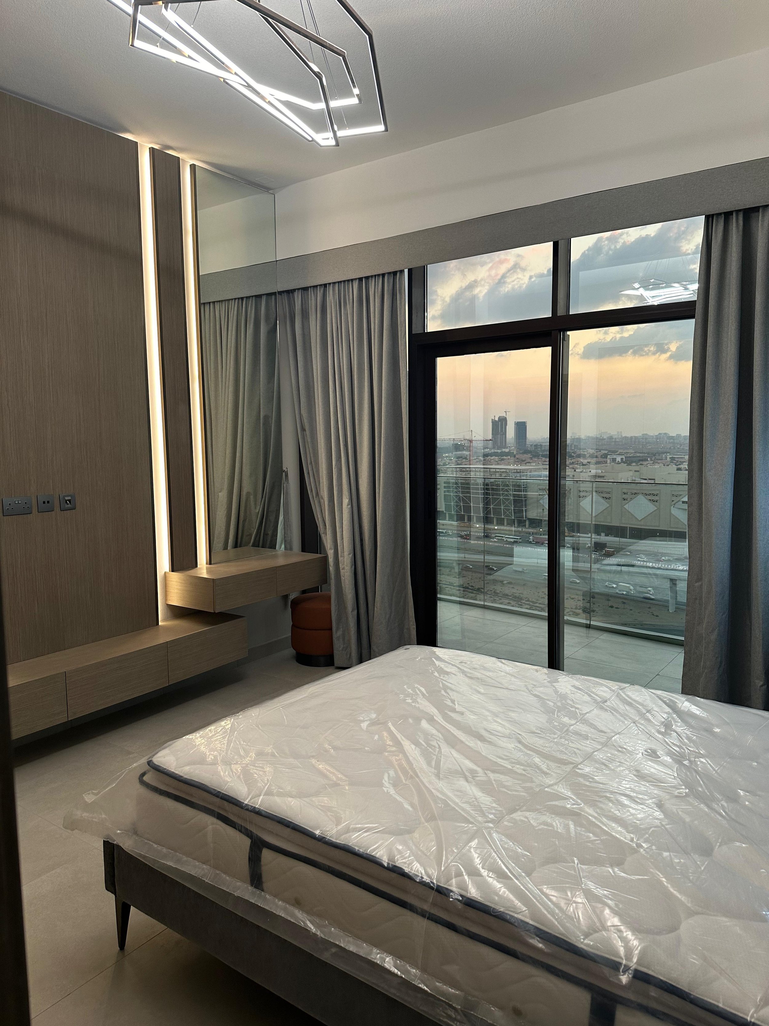 A modern hotel room with a bed covered in plastic, a wall-mounted wooden desk, a round orange stool, and large windows with gray curtains showing a city skyline at sunset.