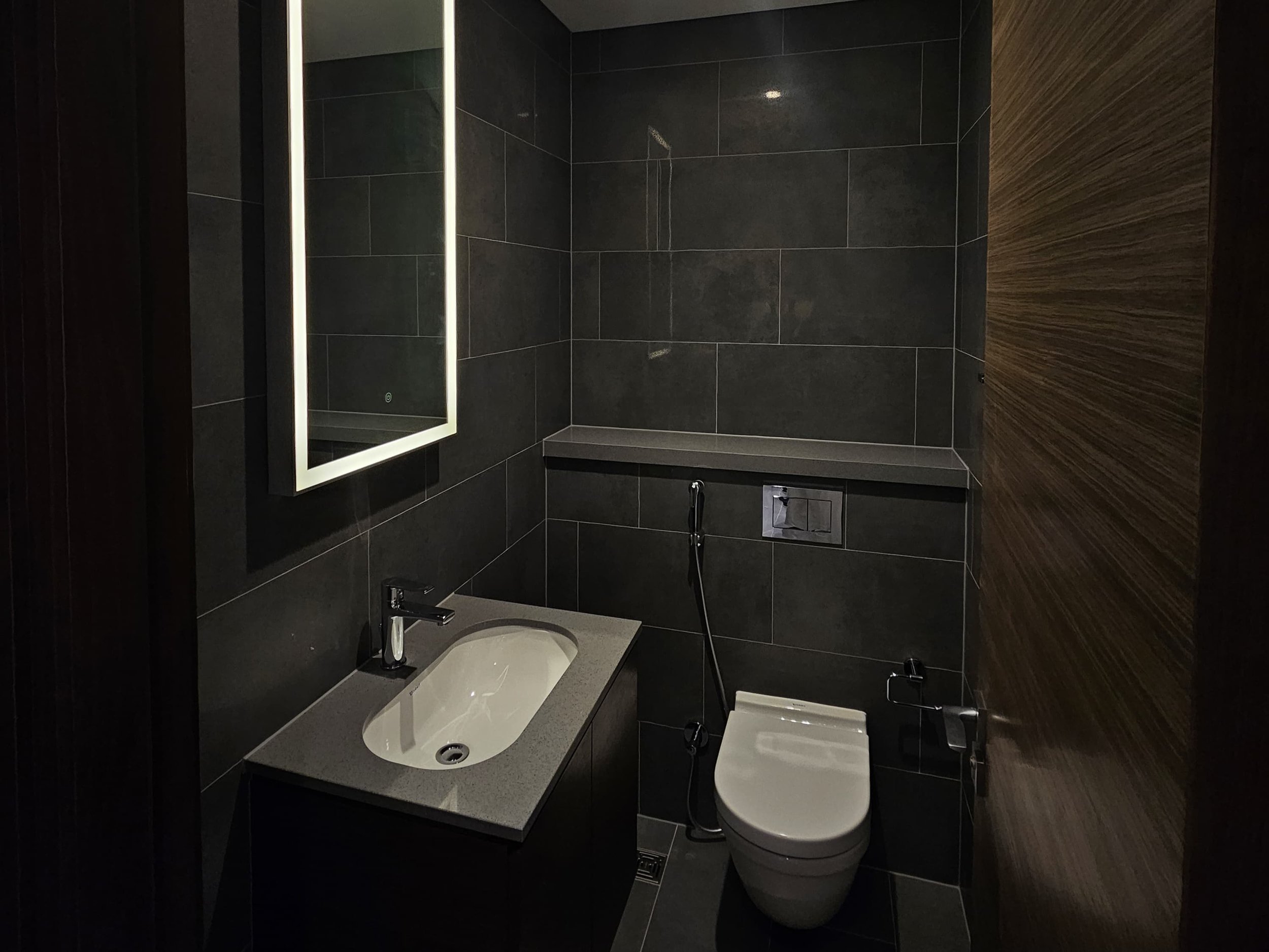 Small modern bathroom with dark gray tiled walls, a wall-mounted mirror with LED light, a small sink with a chrome faucet, and a toilet with a bidet attachment. Brown wood paneling on one side.