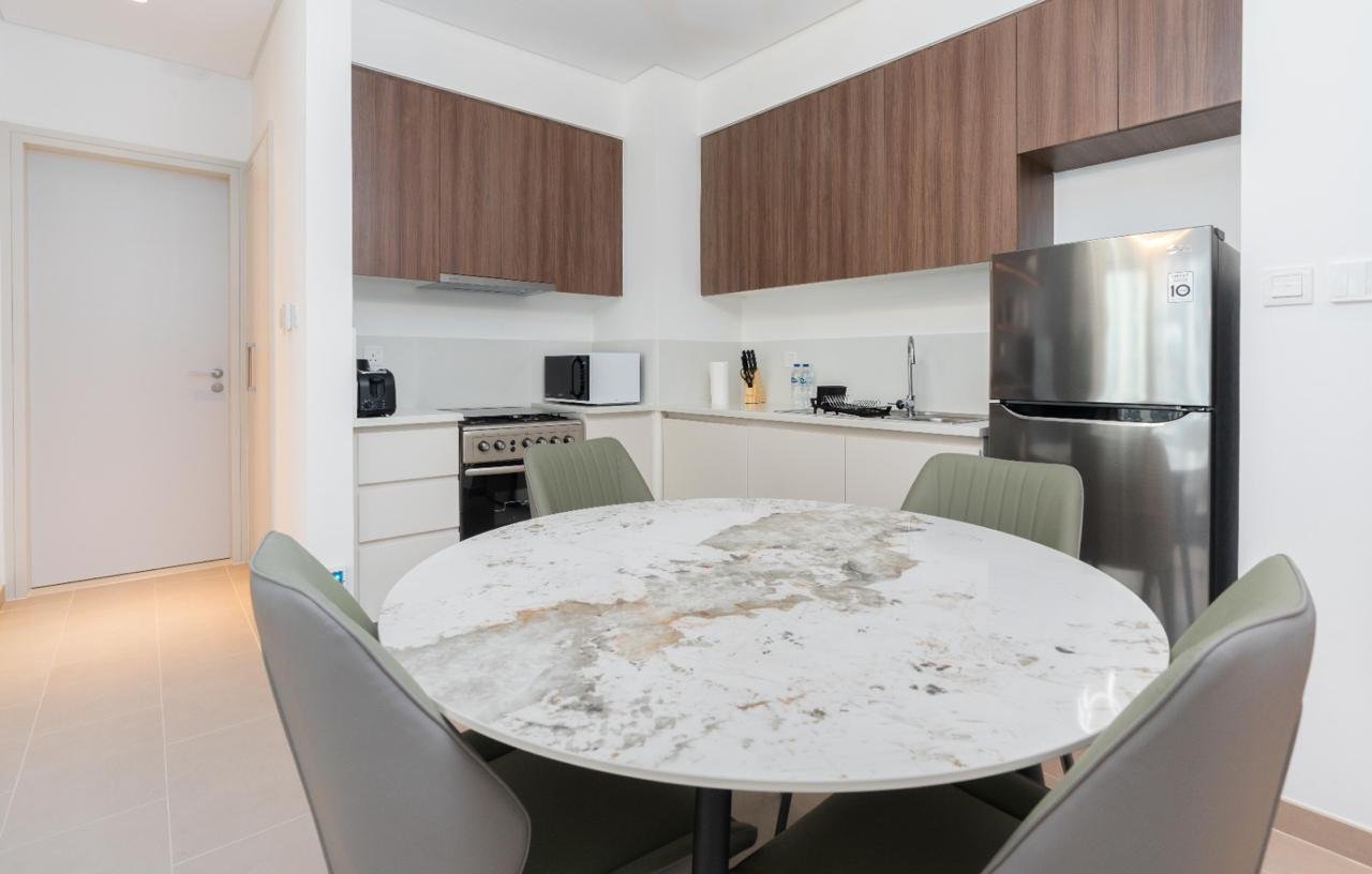 Modern kitchen with a round marble dining table and four beige chairs, white cabinets, wooden upper cabinets, stainless steel refrigerator, microwave, toaster, water bottles, knife block, and a small sink.