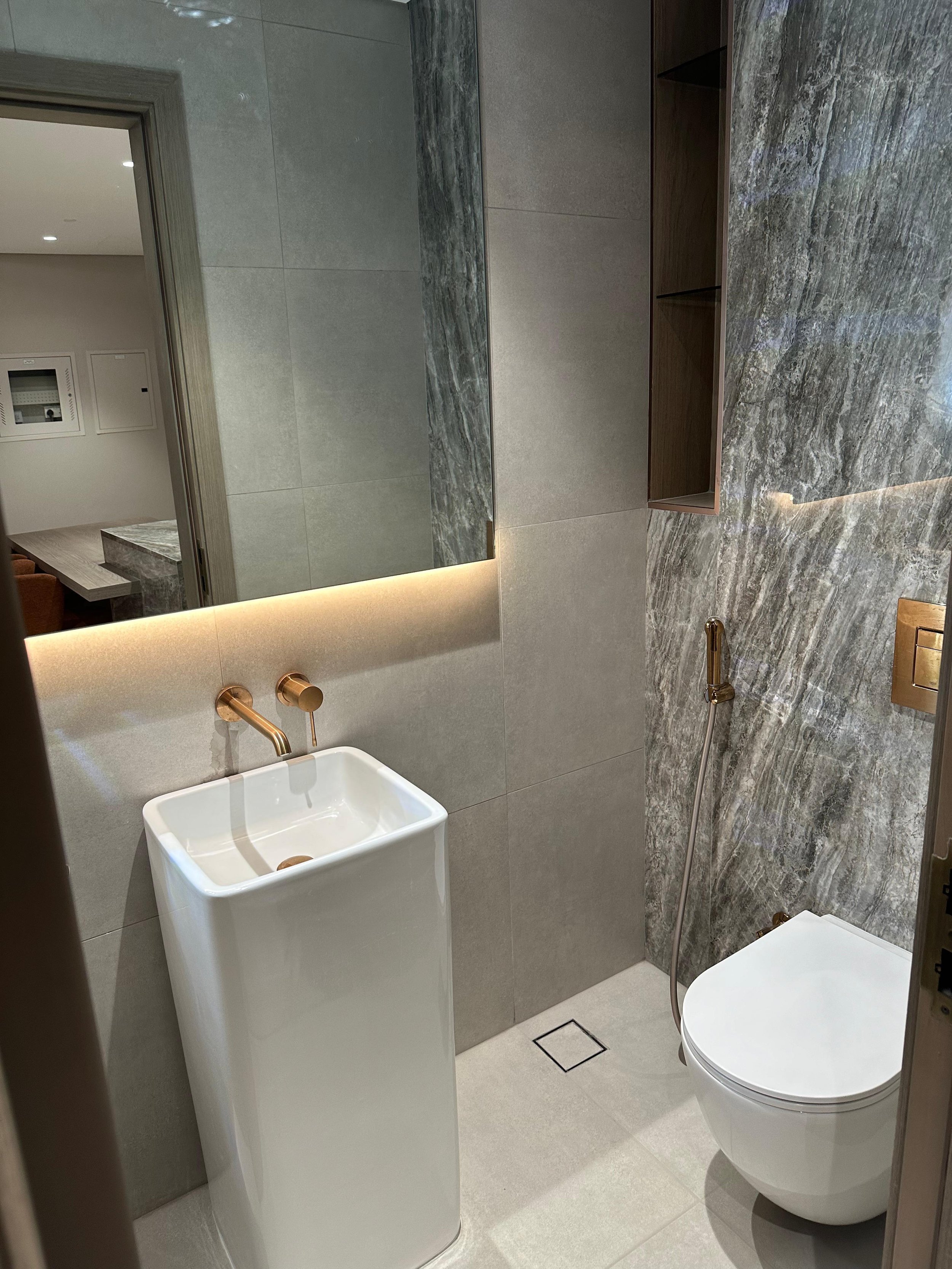 Modern bathroom with a white rectangular sink, a large mirror, a wall-mounted faucet, a toilet with a concealed flush, a handheld bidet sprayer, and gray marble wall tiles.