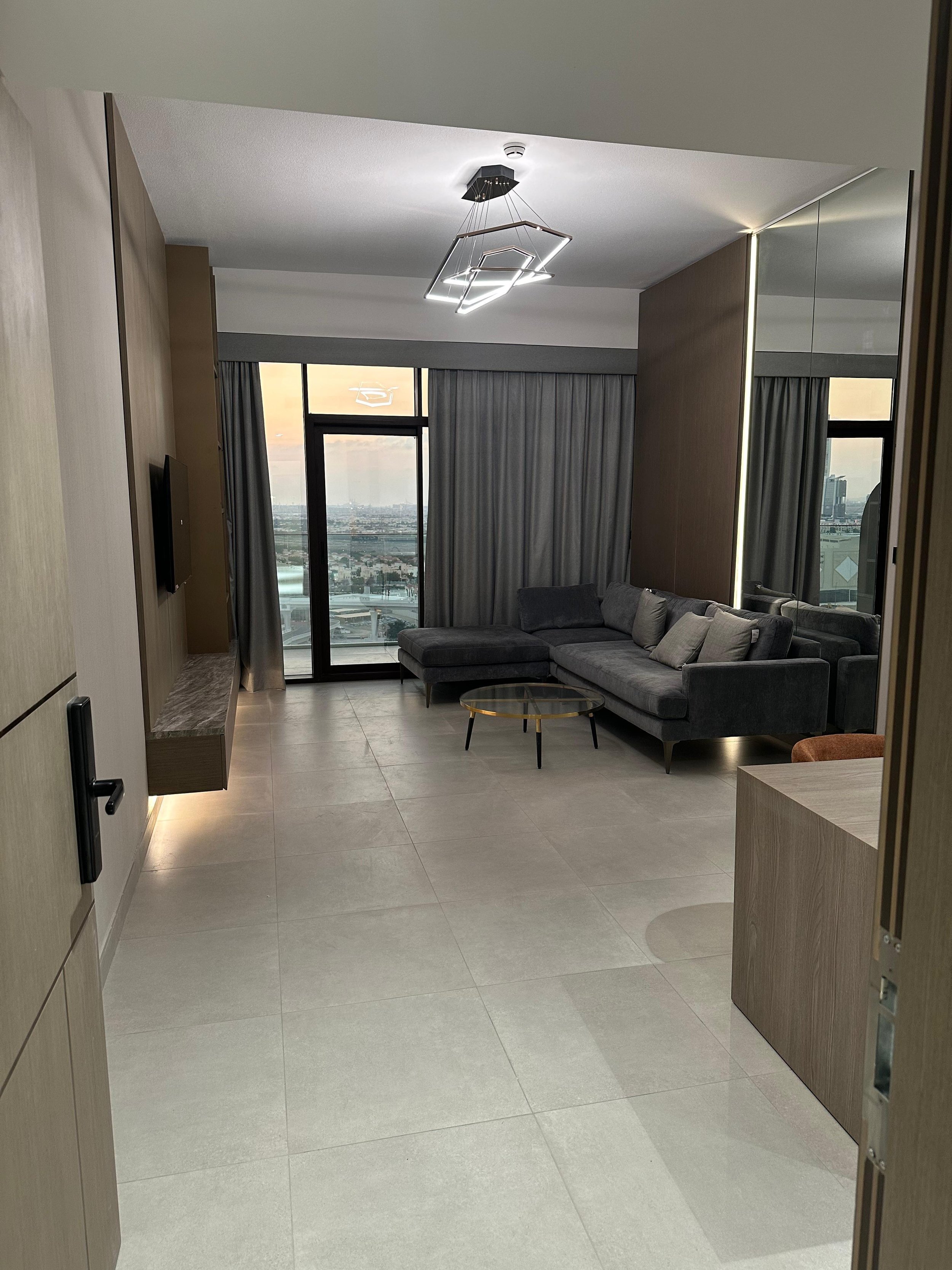 Modern living room with gray sectional sofa, glass coffee table, wall-mounted TV, large window with city view, light-colored tile floor, ceiling light fixture, full-length mirror, and curtains.