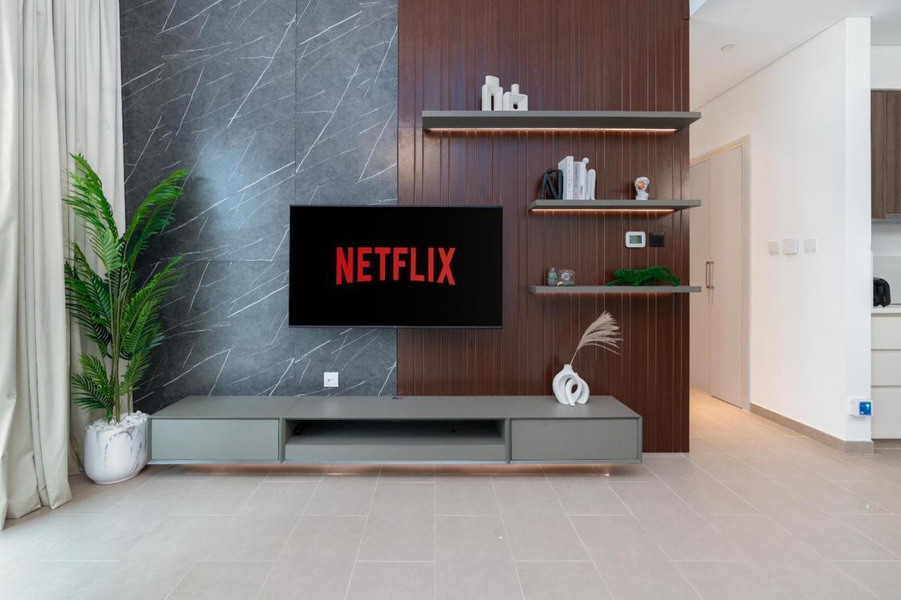 Living room with flat-screen TV displaying Netflix logo, gray TV stand, wooden wall with open shelves, potted green plant, decorative white vase, and part of the kitchen area