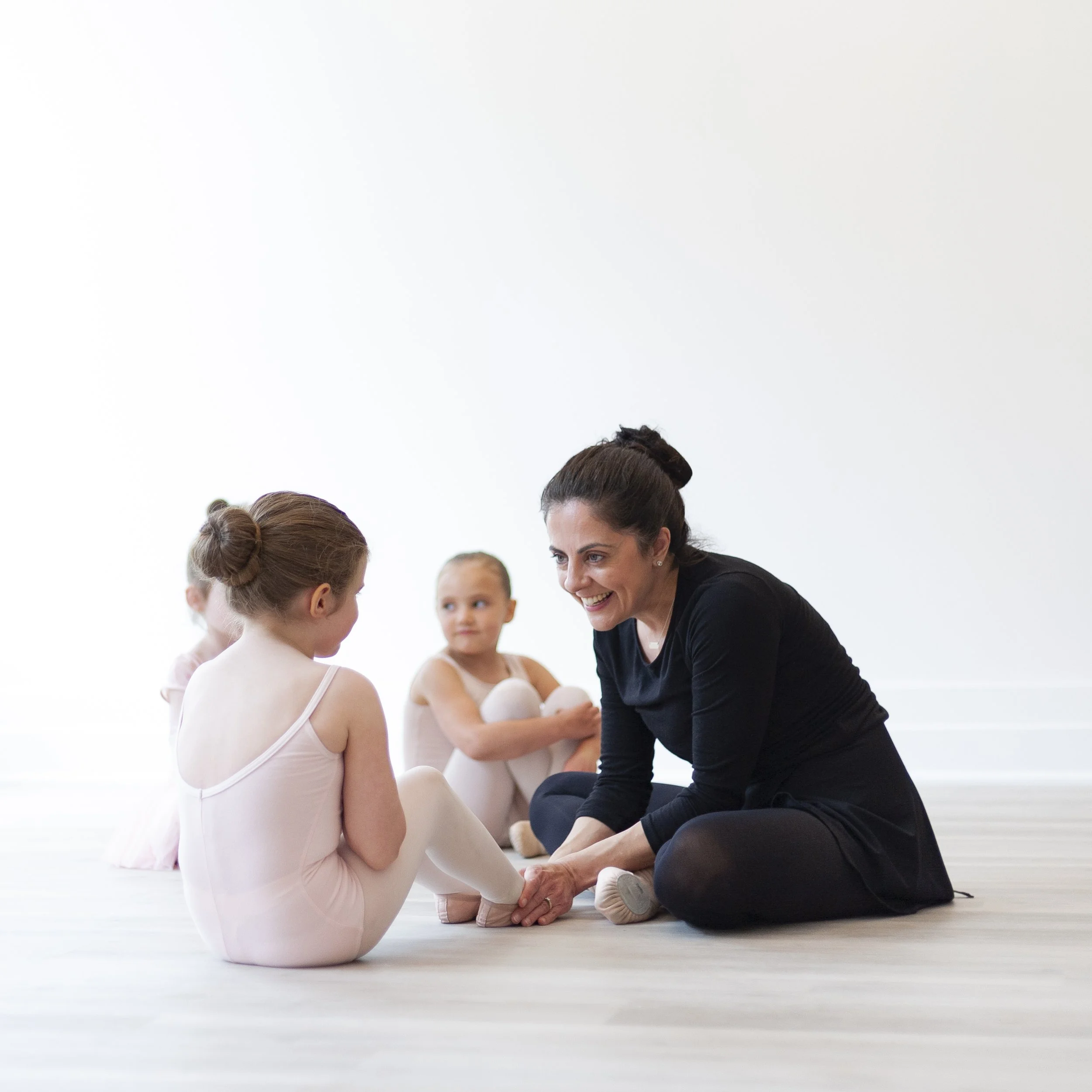 A female ballet instructor sitting on the floor and smiling while talking to a young girl in a ballet outfit, with two other young girls in ballet outfits sitting in the background.