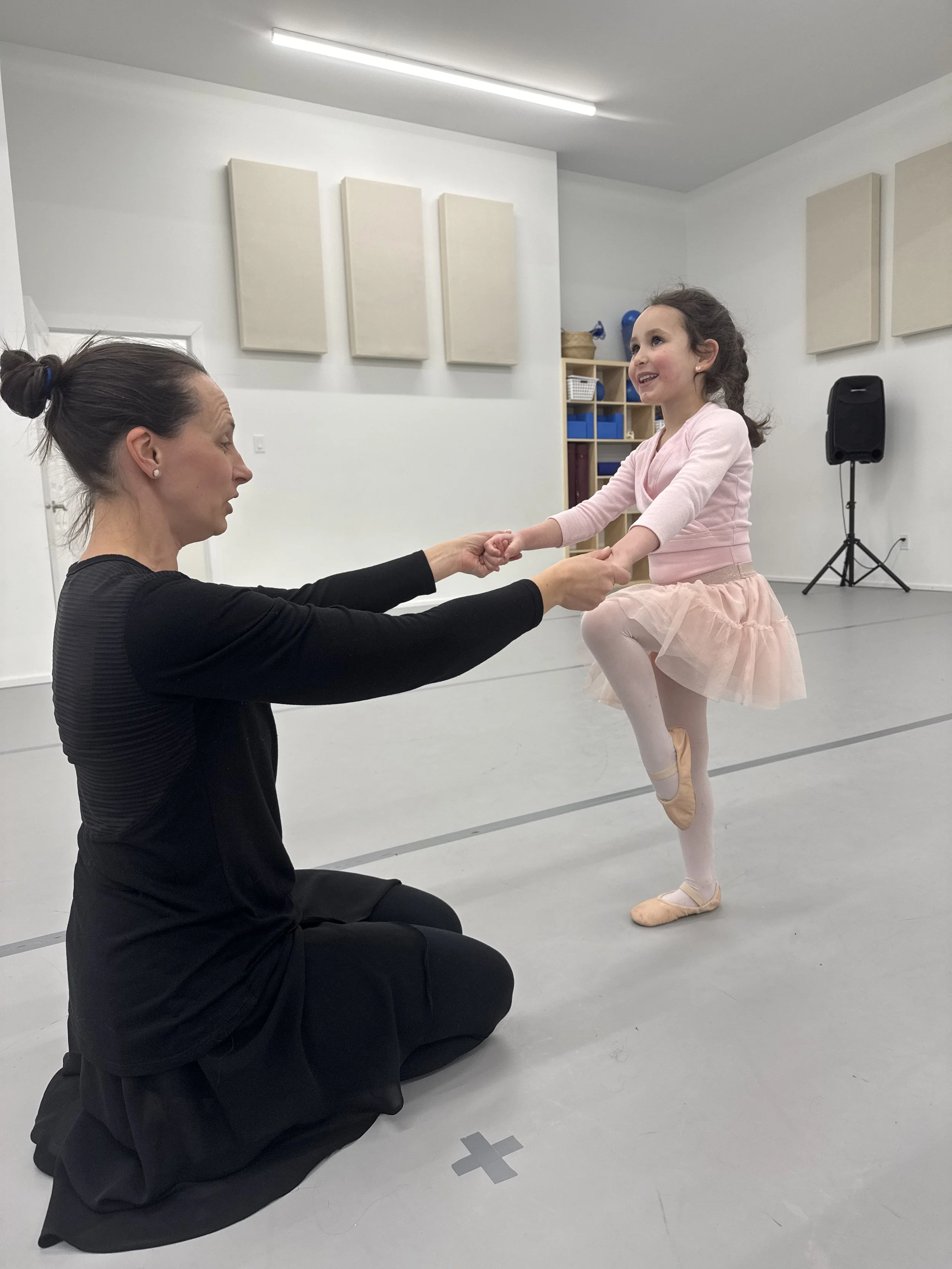 A ballet teacher kneeling on the floor holding hands with a young girl in a pink ballet tutu, who is standing on one leg in a ballet pose, smiling in a dance studio.