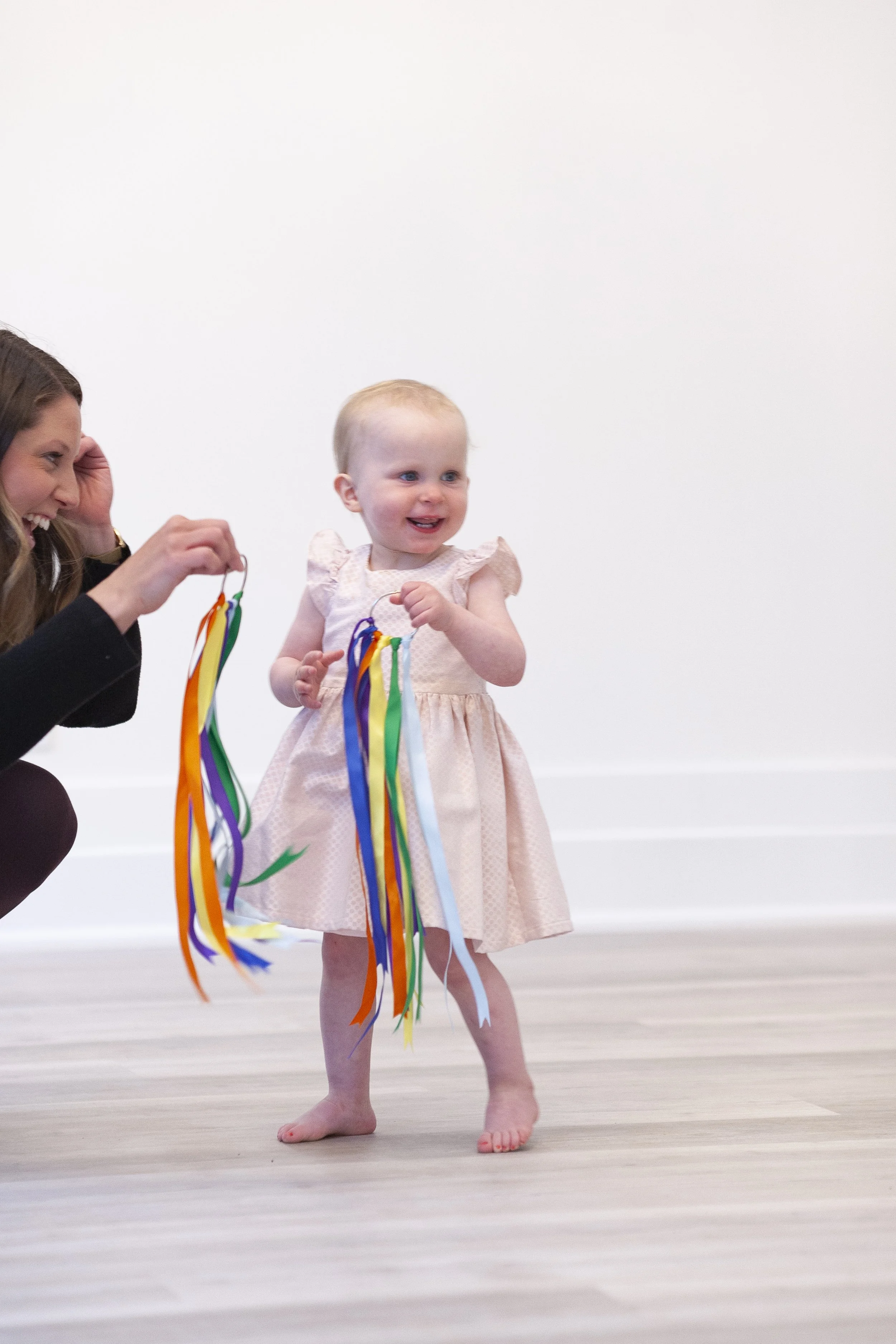 A young girl holding colorful ribbon streamers, smiling, with a woman crouching nearby holding more streamers and smiling.