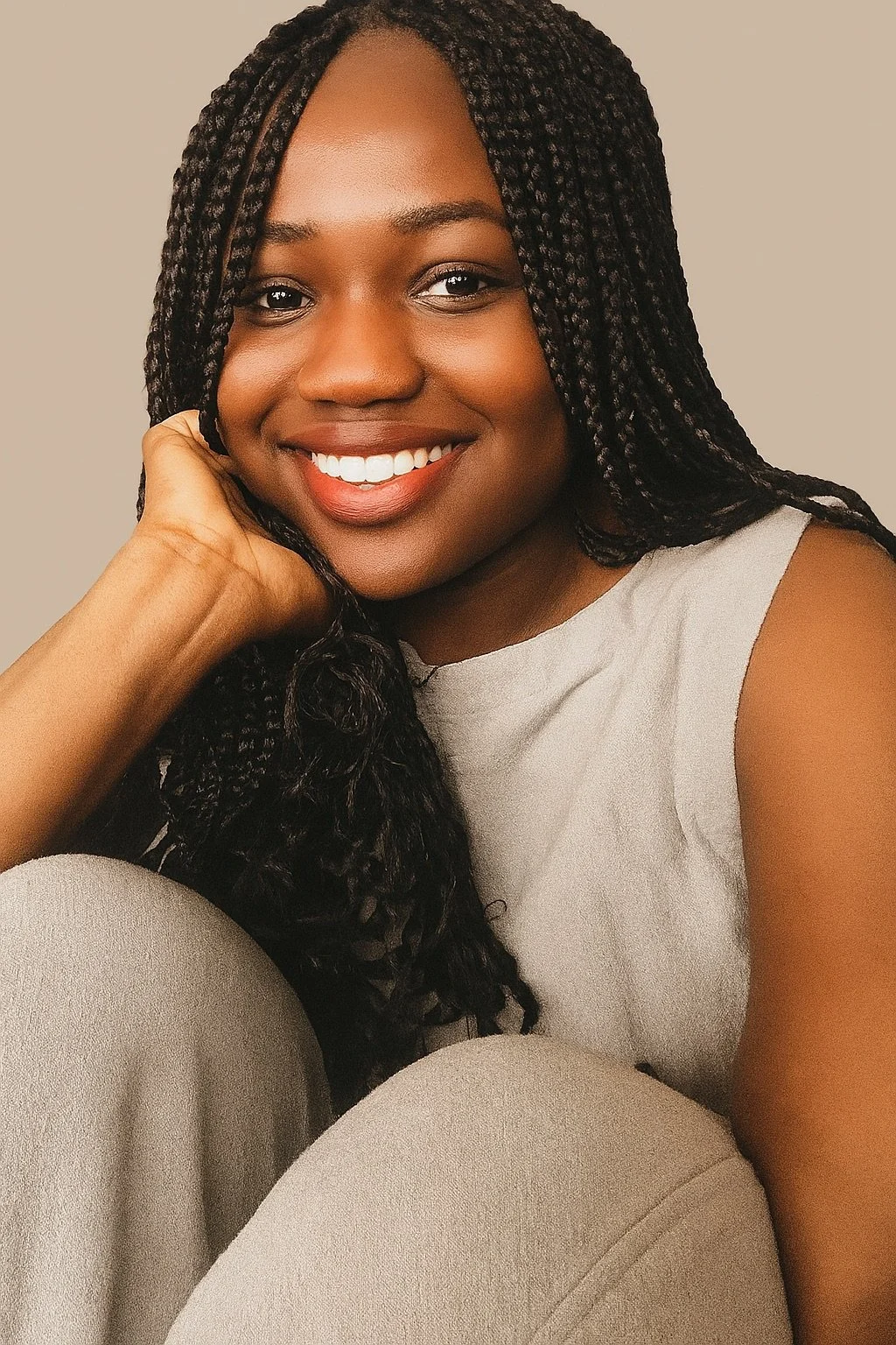 A young woman with dark braids, smiling, resting her face on her hand, wearing a light-colored sleeveless top, sitting against a neutral background.