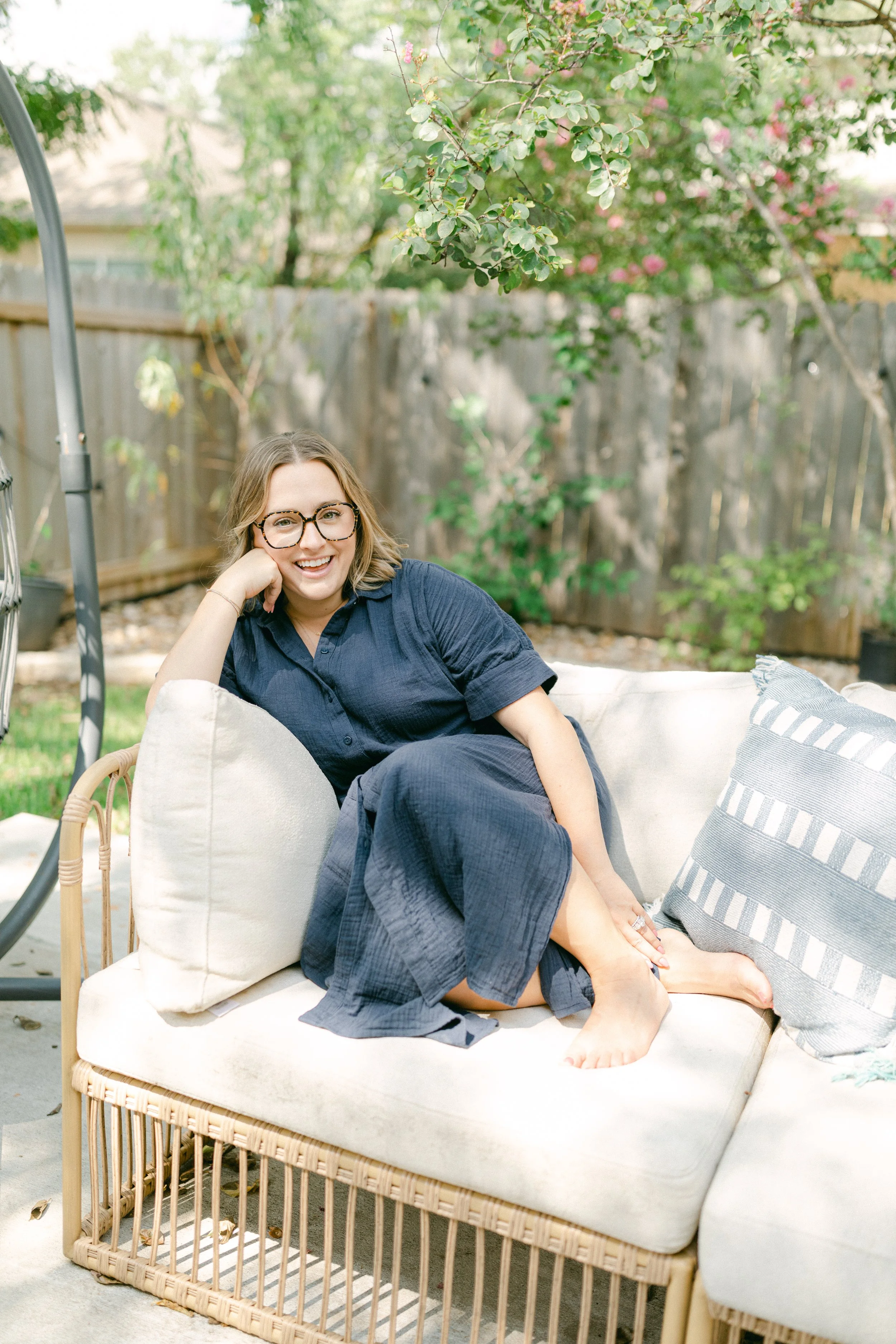 A woman with glasses and a navy blue dress relaxing on an outdoor rattan sofa with a white cushion and striped pillows in a garden with a wooden fence and flowering trees.