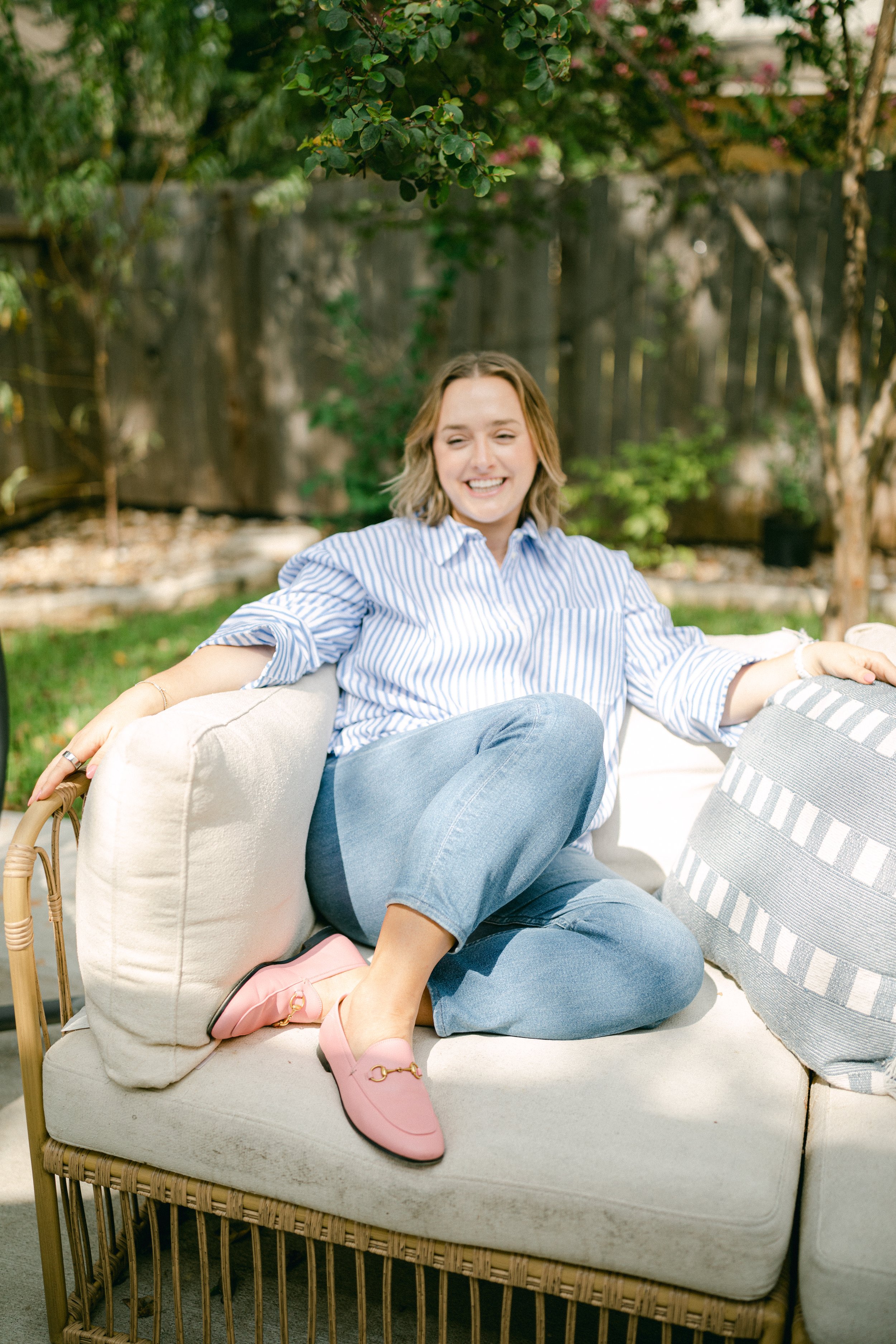 A young woman wearing a blue and white striped shirt, blue jeans, and pink loafers sitting on an outdoor sofa, smiling and relaxing in a garden setting.