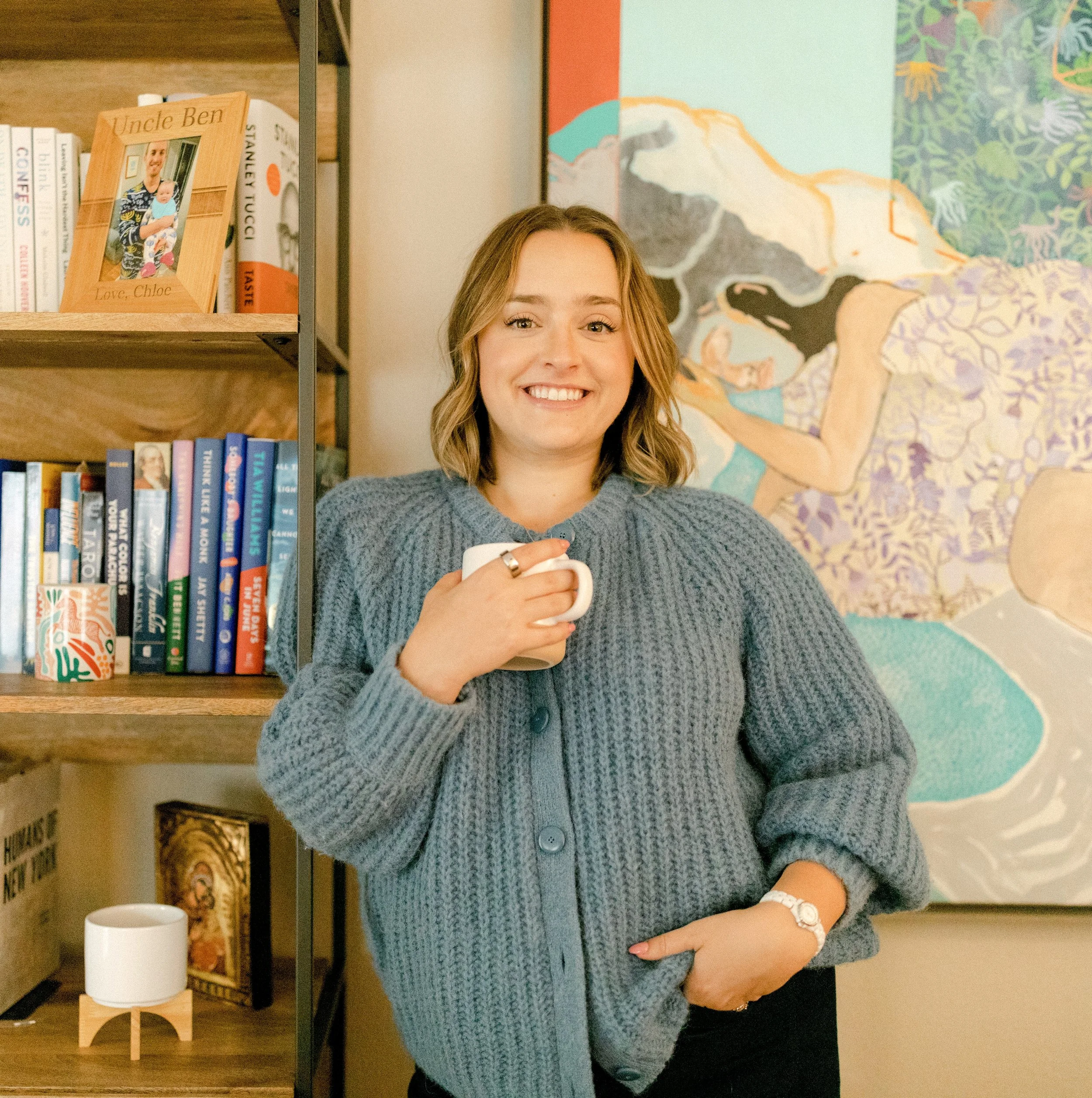 A woman with shoulder-length wavy blonde hair smiling and holding a white mug, standing in front of a bookshelf and colorful artwork.