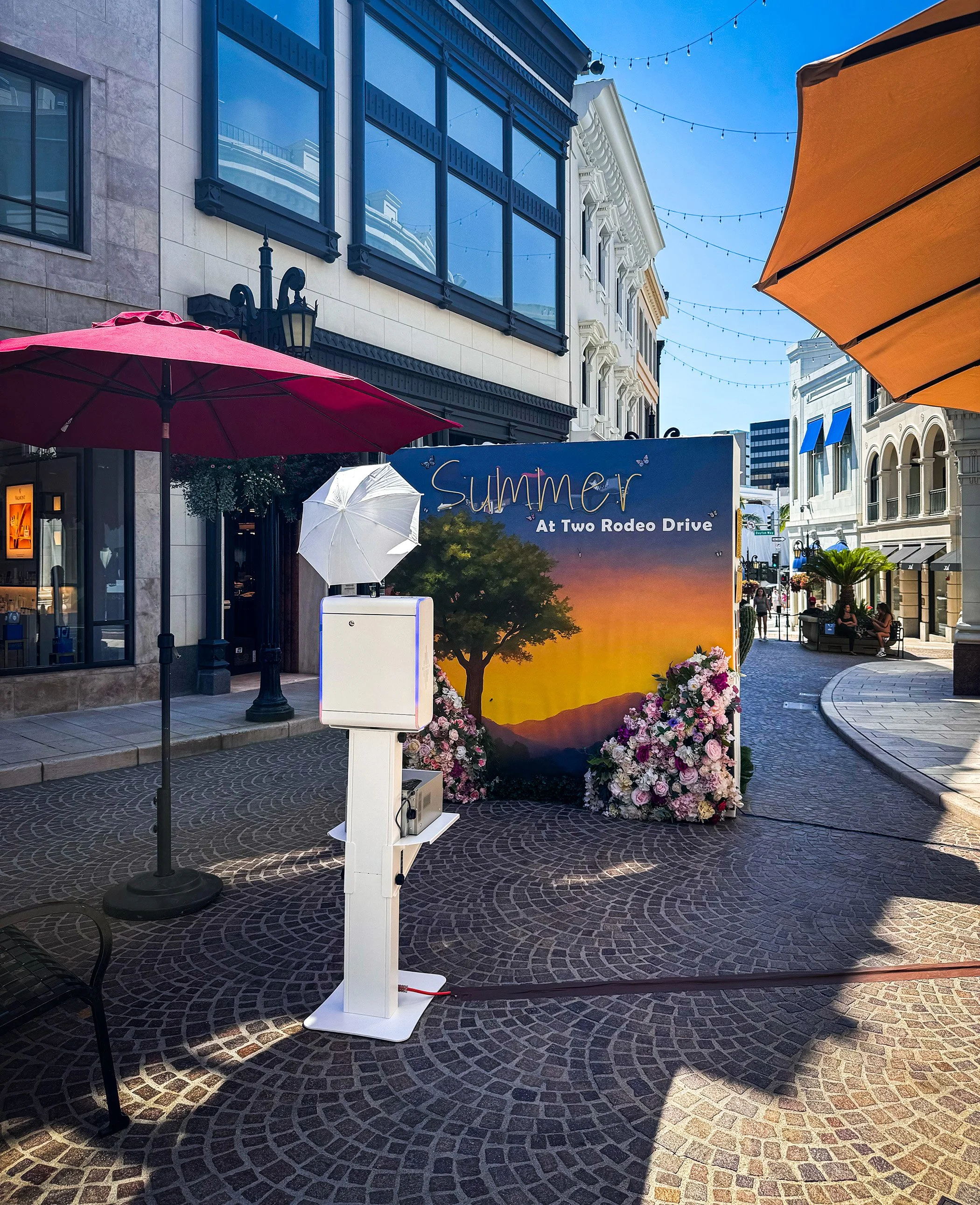 Outdoor shopping mall with storefronts, umbrellas, and a promotional backdrop for summer at Two Rodeo Drive, featuring flowers and a sunset scene.