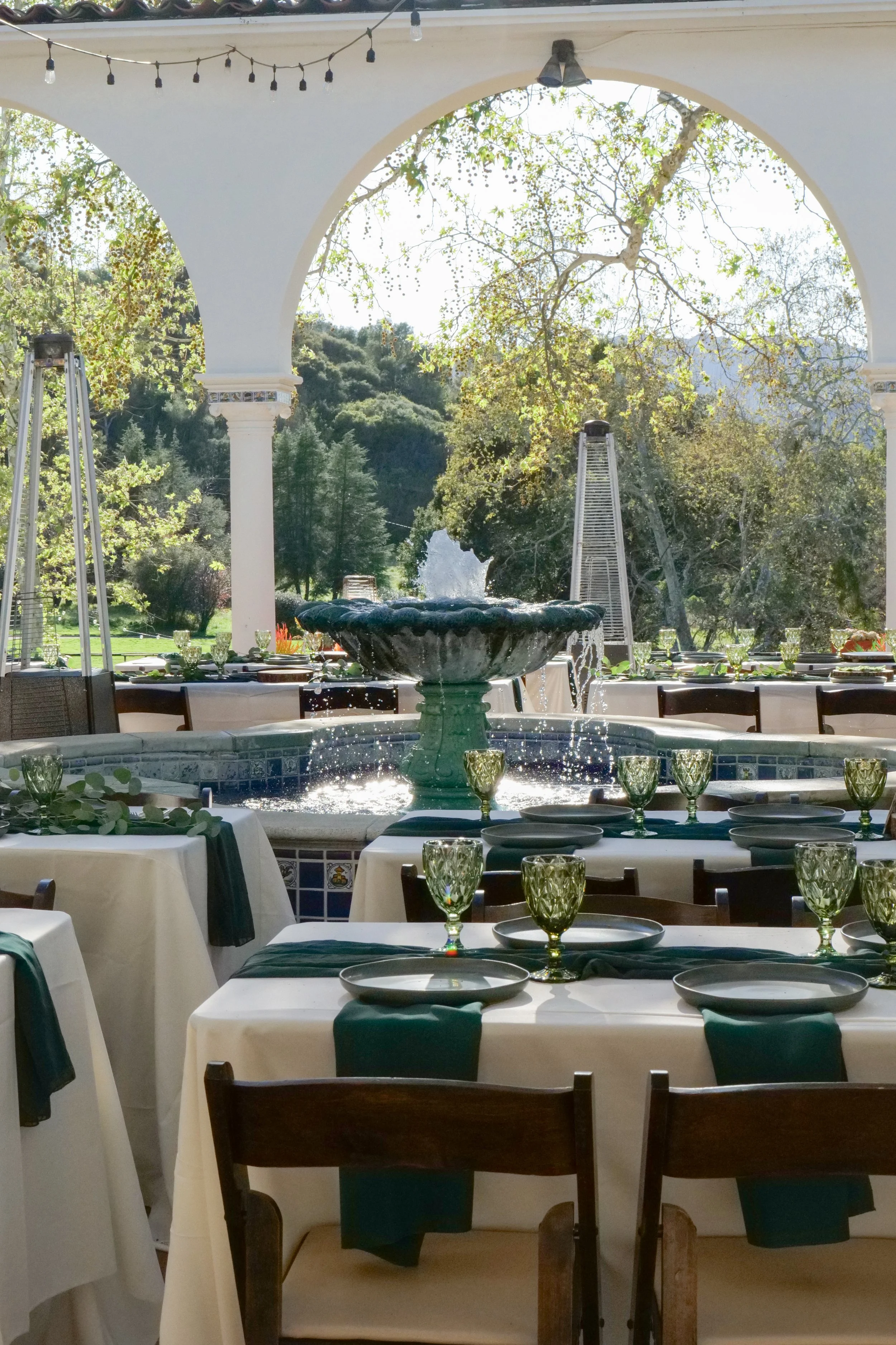 Indoor dining setup with tables covered in white tablecloths, black napkins, and green glasses, in front of large arched windows showing trees and a fountain outside, with string lights hanging from the ceiling.