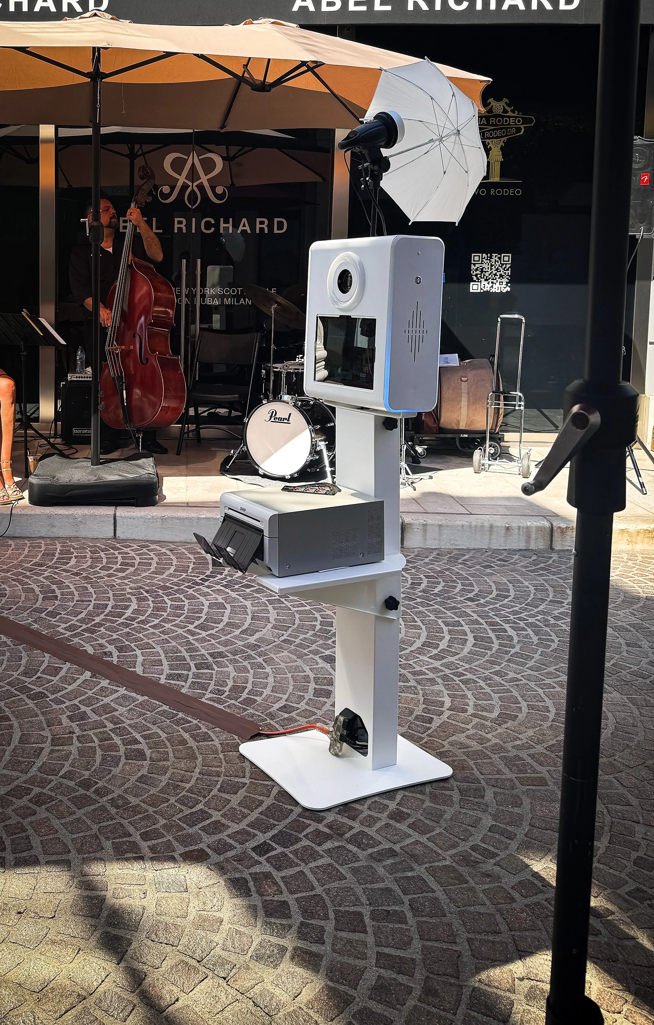 Photo of a photo booth set up outdoors on a brick sidewalk, with a band playing on a stage behind it, including a double bass, drums, and a keyboard, under an umbrella.
