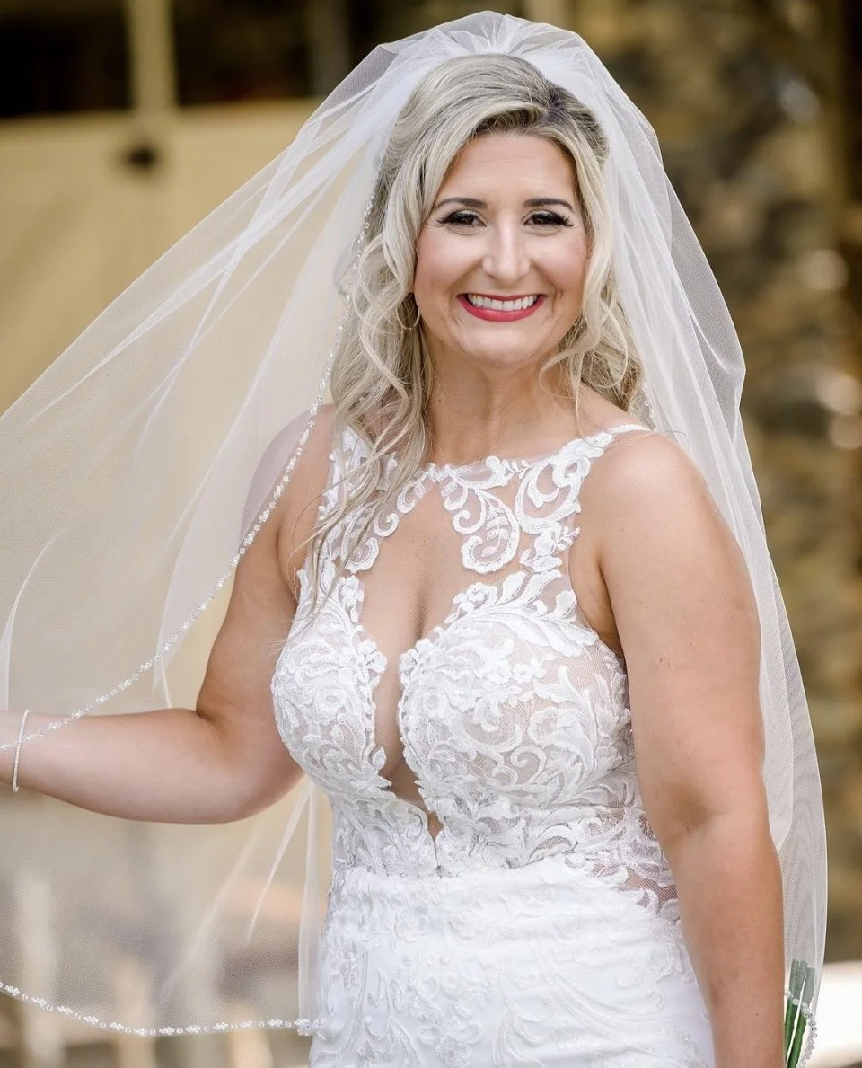 A smiling bride in a white lace wedding dress with a veil, outdoors.