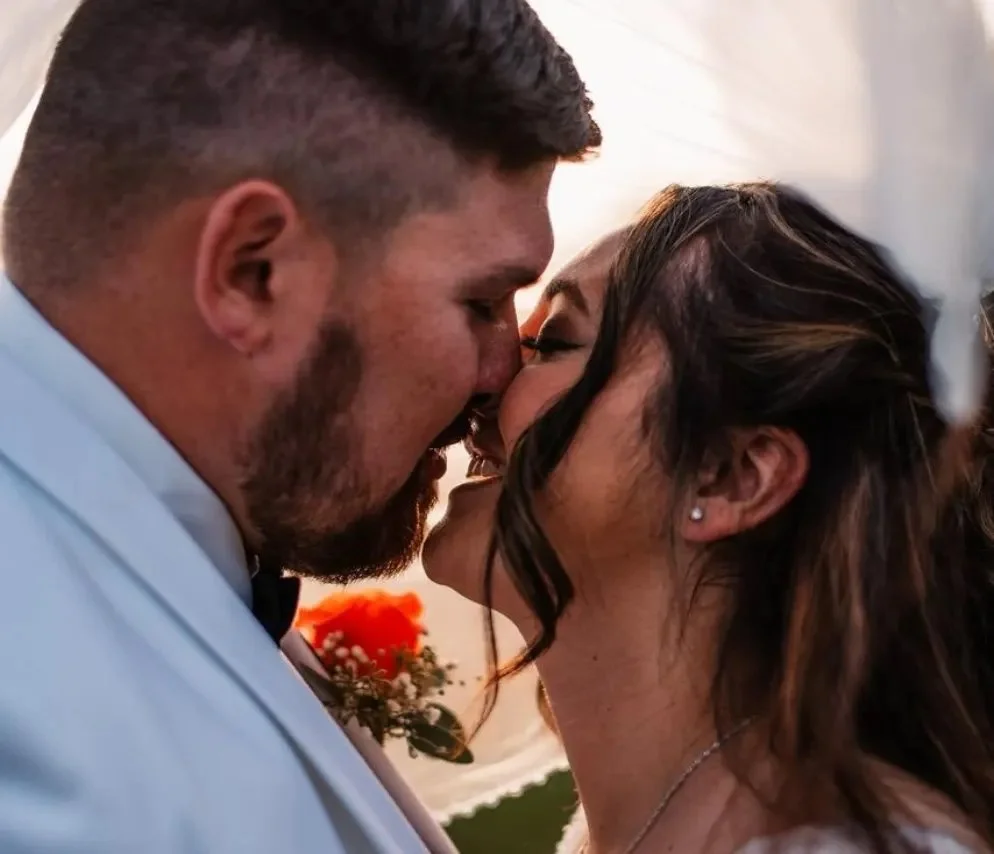 A couple is sharing a kiss at their wedding, with the man wearing a white tuxedo and the woman having dark, styled hair and earrings.