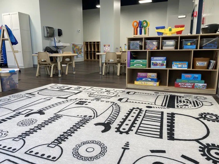 An empty classroom with a large black and white patterned rug on the floor, small tables and chairs, and shelves filled with educational toys and supplies.