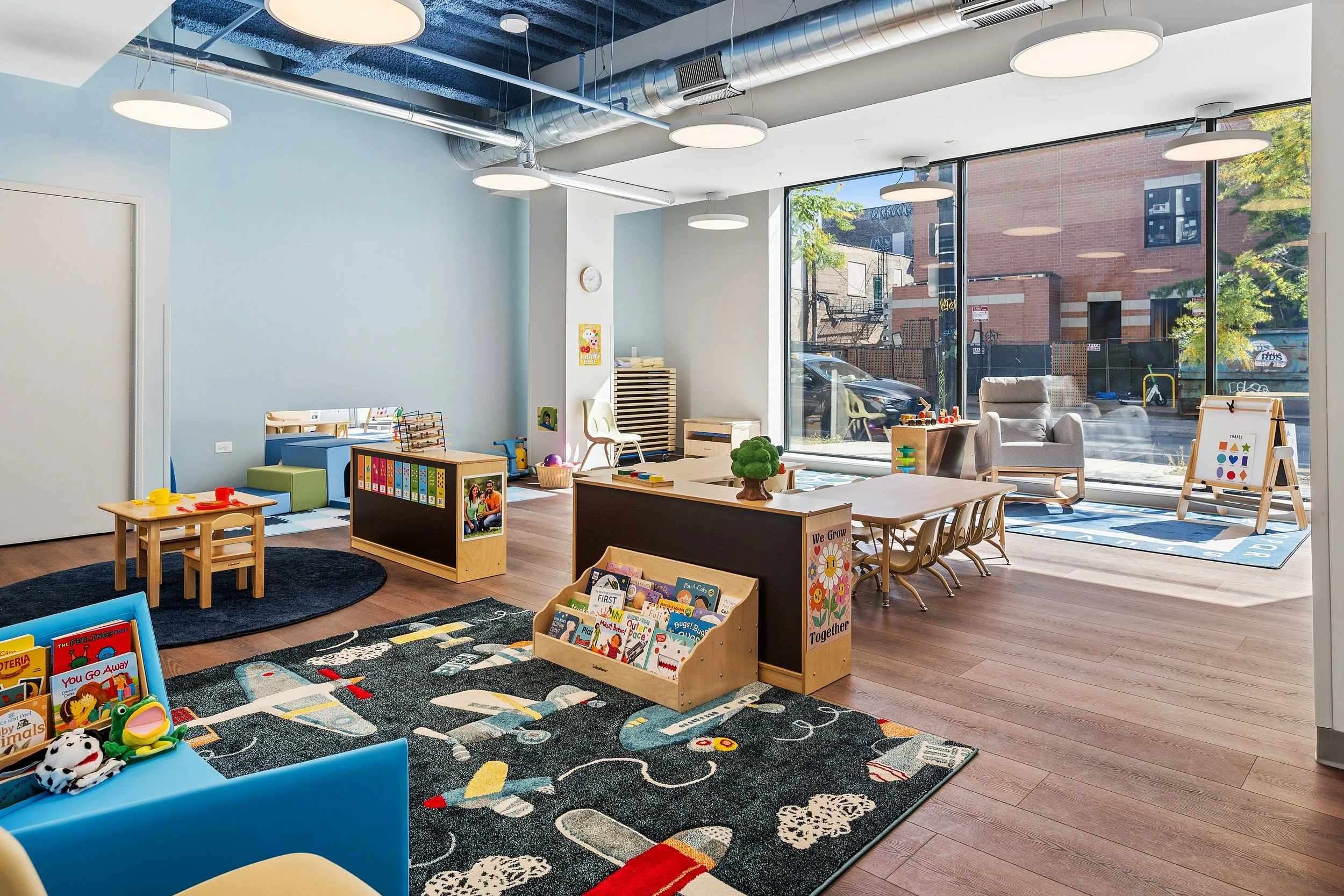 A spacious classroom with multiple small tables and chairs, a colorful rug, and shelves with educational materials, illuminated by large windows.