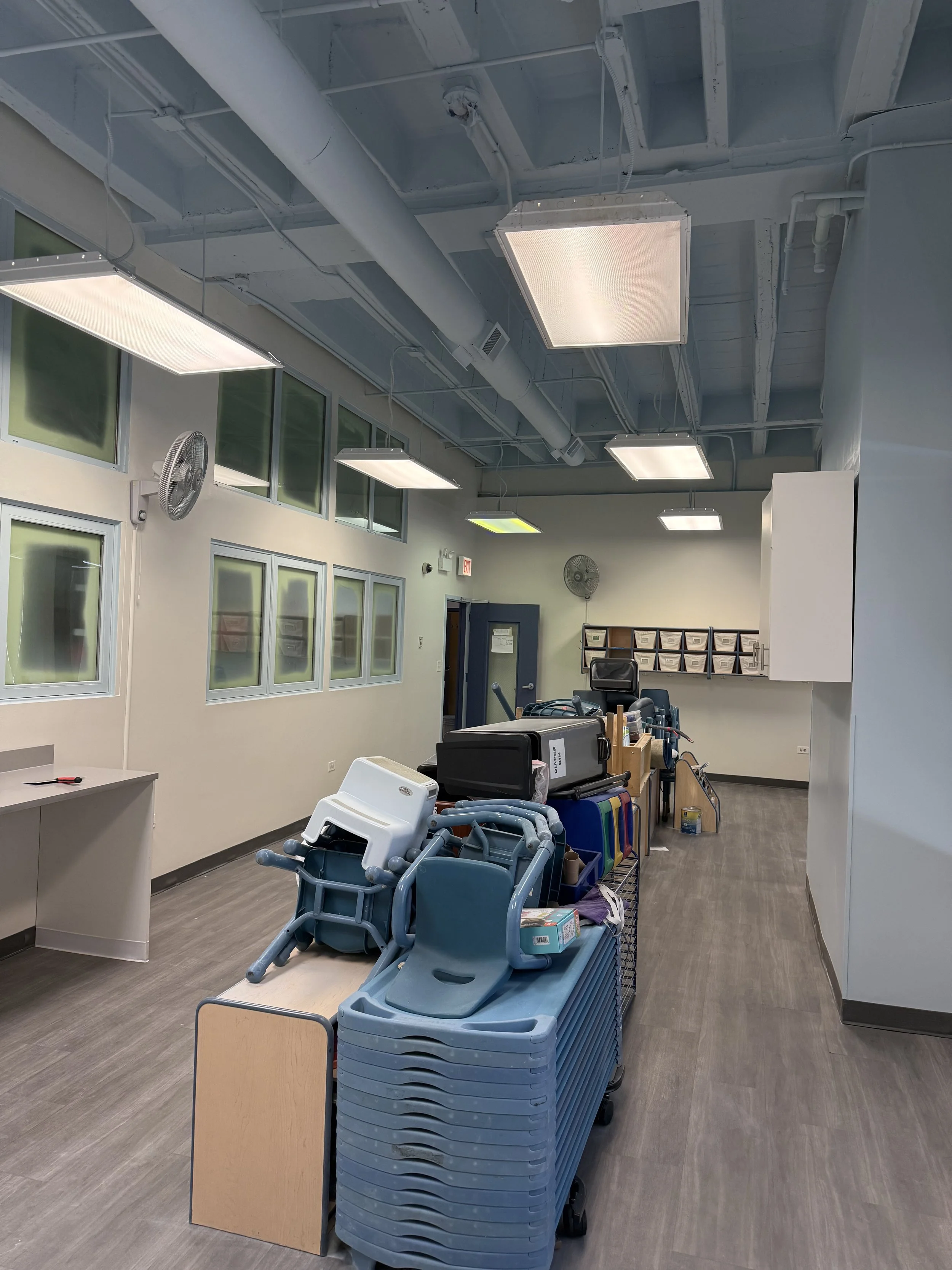 Woman standing on city sidewalk in front of a beige brick building with large windows, smiling, wearing a brown jacket and black pants.