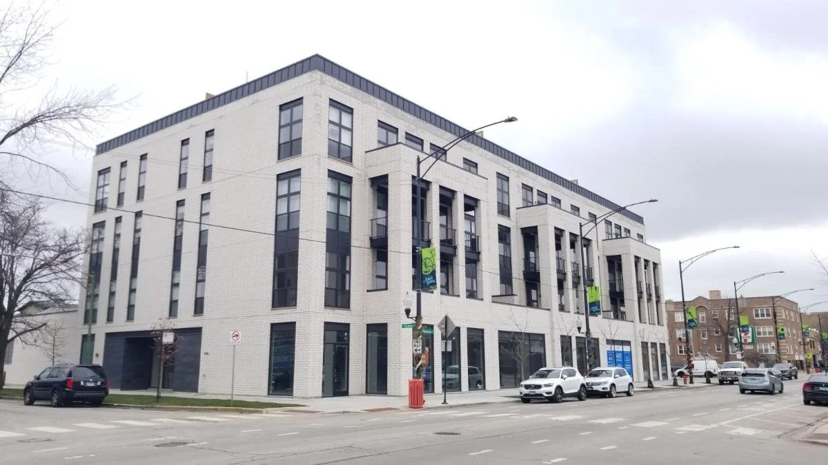 Modern white brick building with large windows, situated on a city street with parked cars, streetlights, and a cloudy sky.