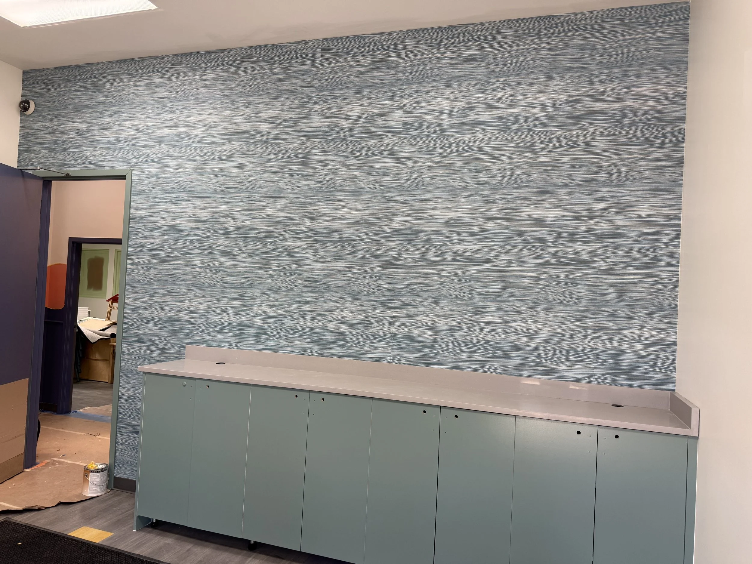 A spacious classroom with multiple small tables and chairs, a colorful rug, and shelves with educational materials, illuminated by large windows.