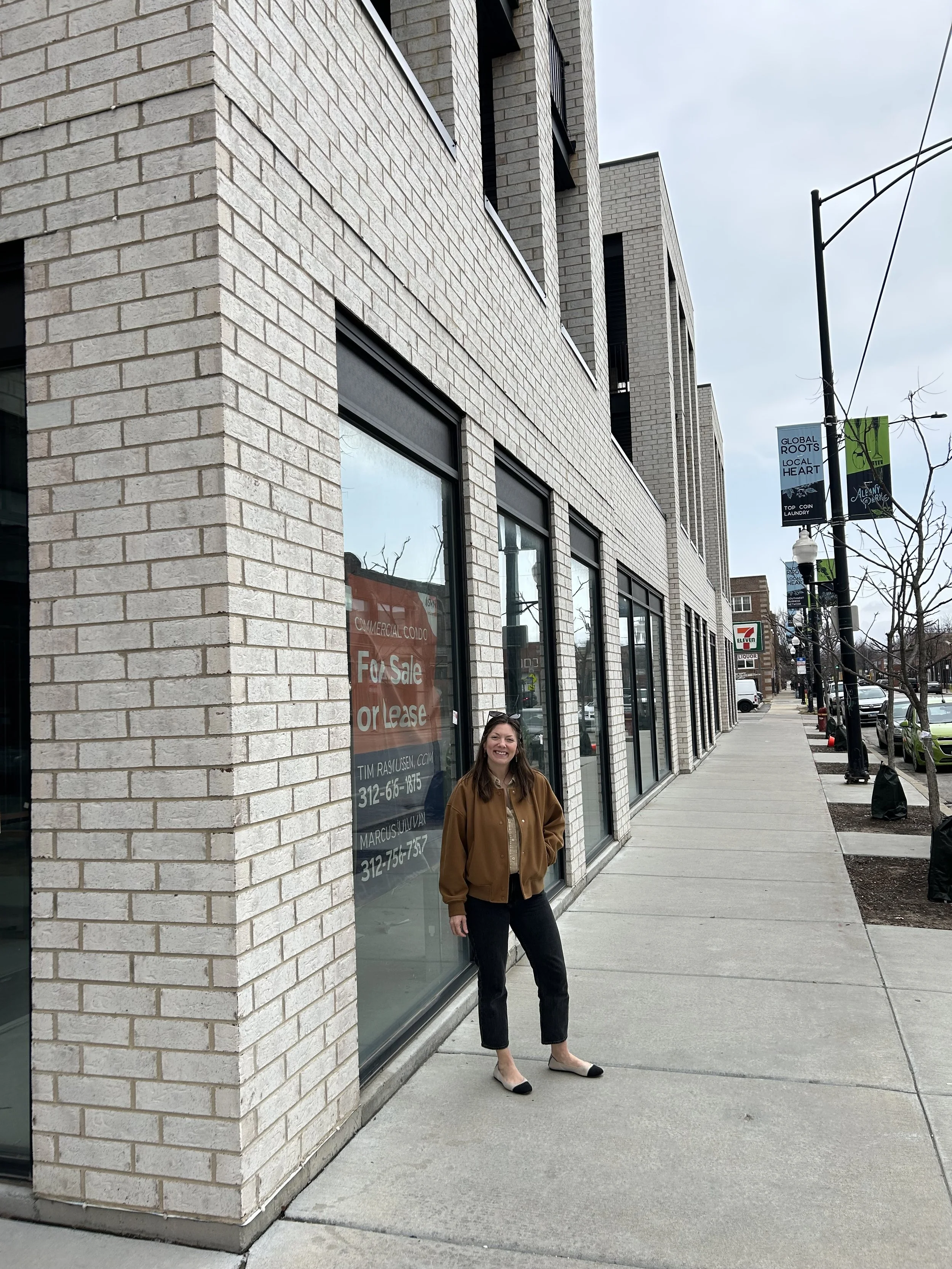 Woman standing on city sidewalk in front of a beige brick building with large windows, smiling, wearing a brown jacket and black pants.