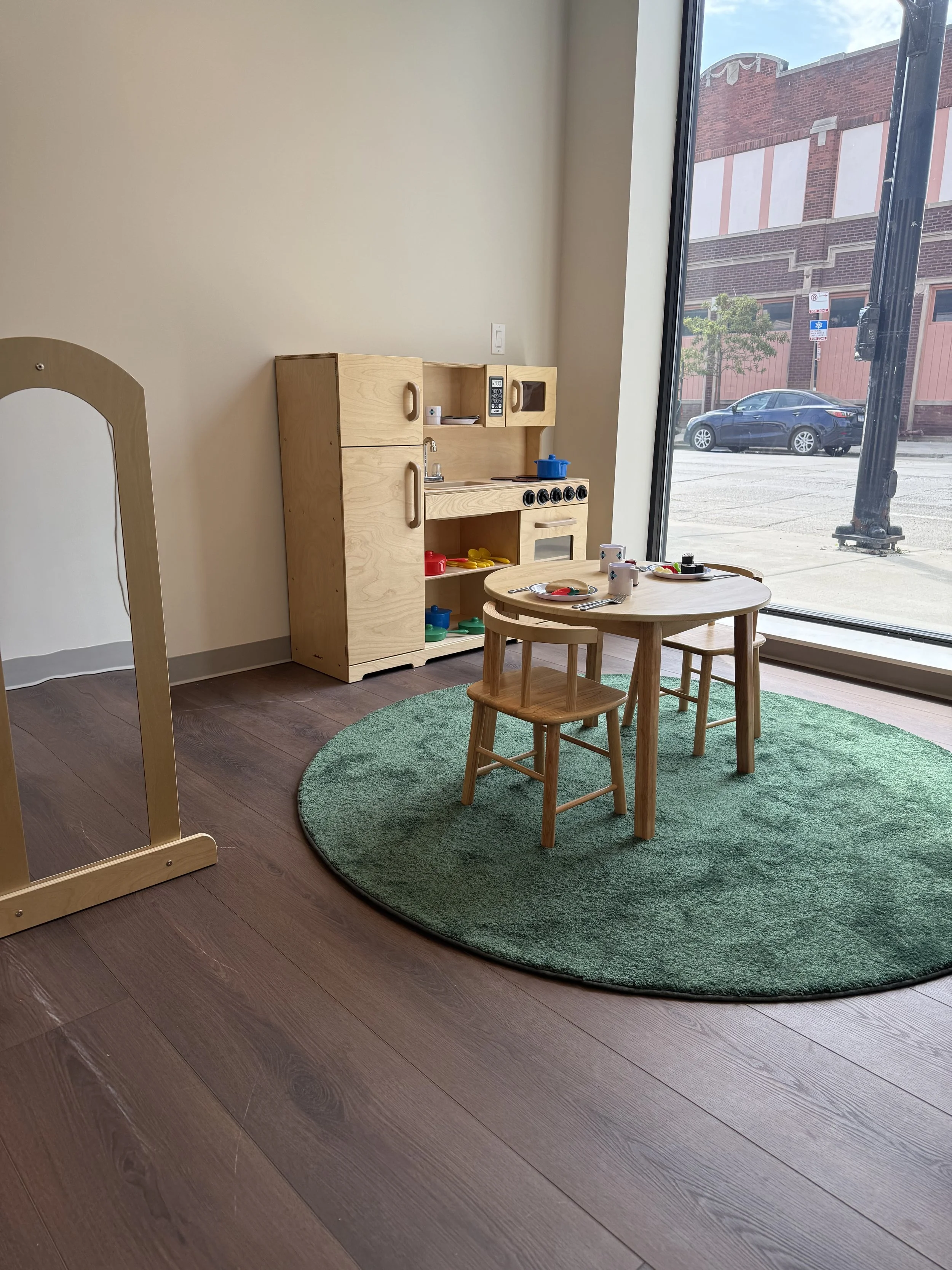 A spacious classroom with multiple small tables and chairs, a colorful rug, and shelves with educational materials, illuminated by large windows.