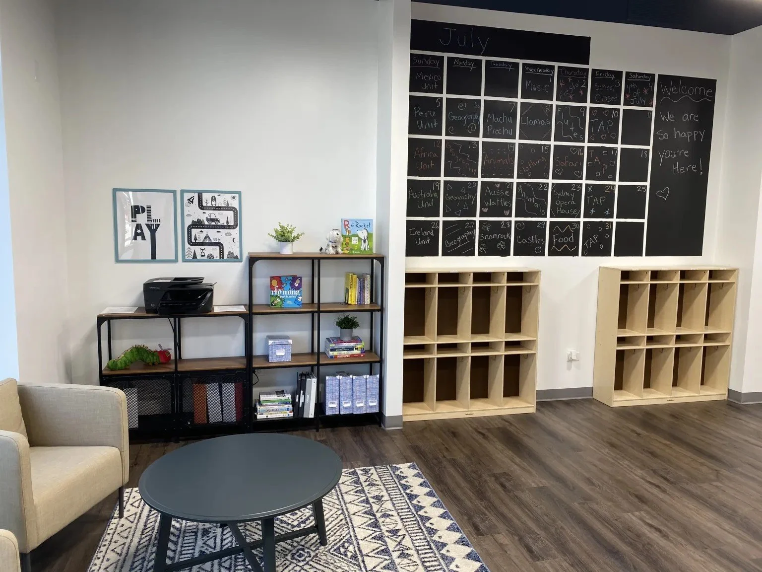 A classroom corner with a black and beige shelving unit, a beige armchair, a round black table, and a large black chalkboard calendar on the wall with July written at the top, featuring various themed activities and notes.