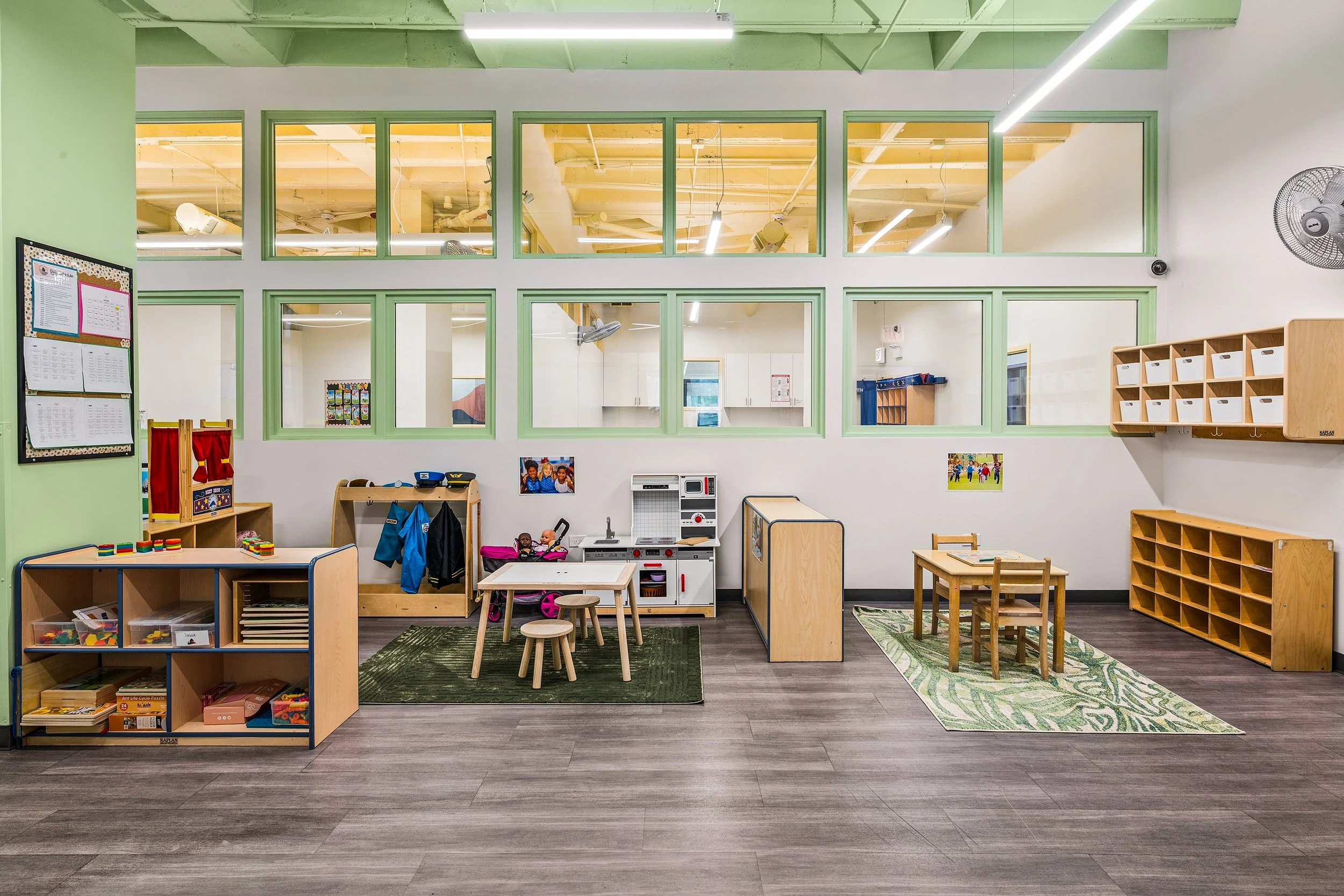 A spacious classroom with multiple small tables and chairs, a colorful rug, and shelves with educational materials, illuminated by large windows.
