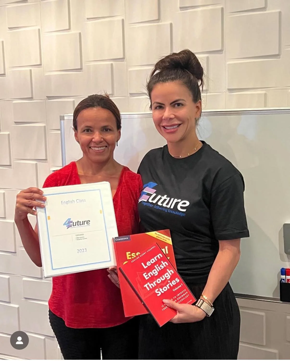 Two women standing together indoors, holding certificates and books. One woman wears a red blouse, the other wears a black T-shirt with 'Future' written on it. They are smiling and standing against a white wall with decorative panels.