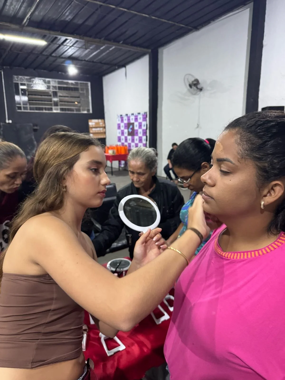 Young woman applying makeup or face paint to another woman in a crowded indoor setting.