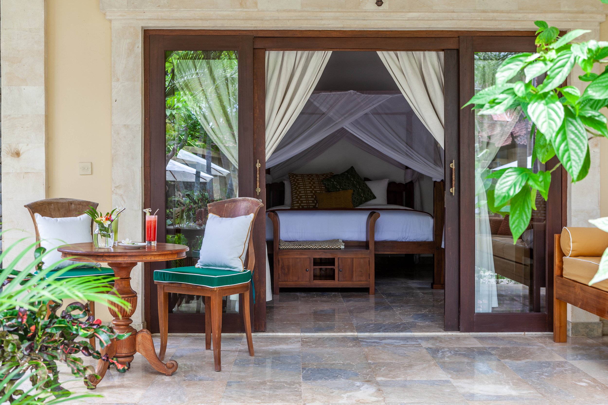 A cozy outdoor patio with a small round wooden table, two wooden chairs with white pillows, a colorful drink, and lush green plants, leading to a bedroom with a canopy bed and cream curtains.