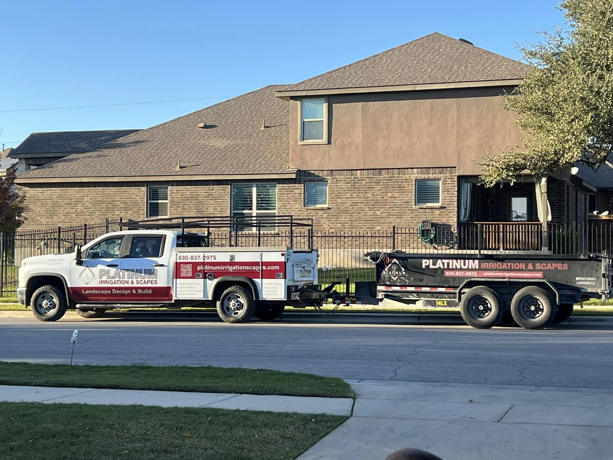 A pickup truck with a trailer parked on the street in front of a residential house. The truck and trailer are branded with "PLATINUM IRRIGATION & SCAPES" advertisements.