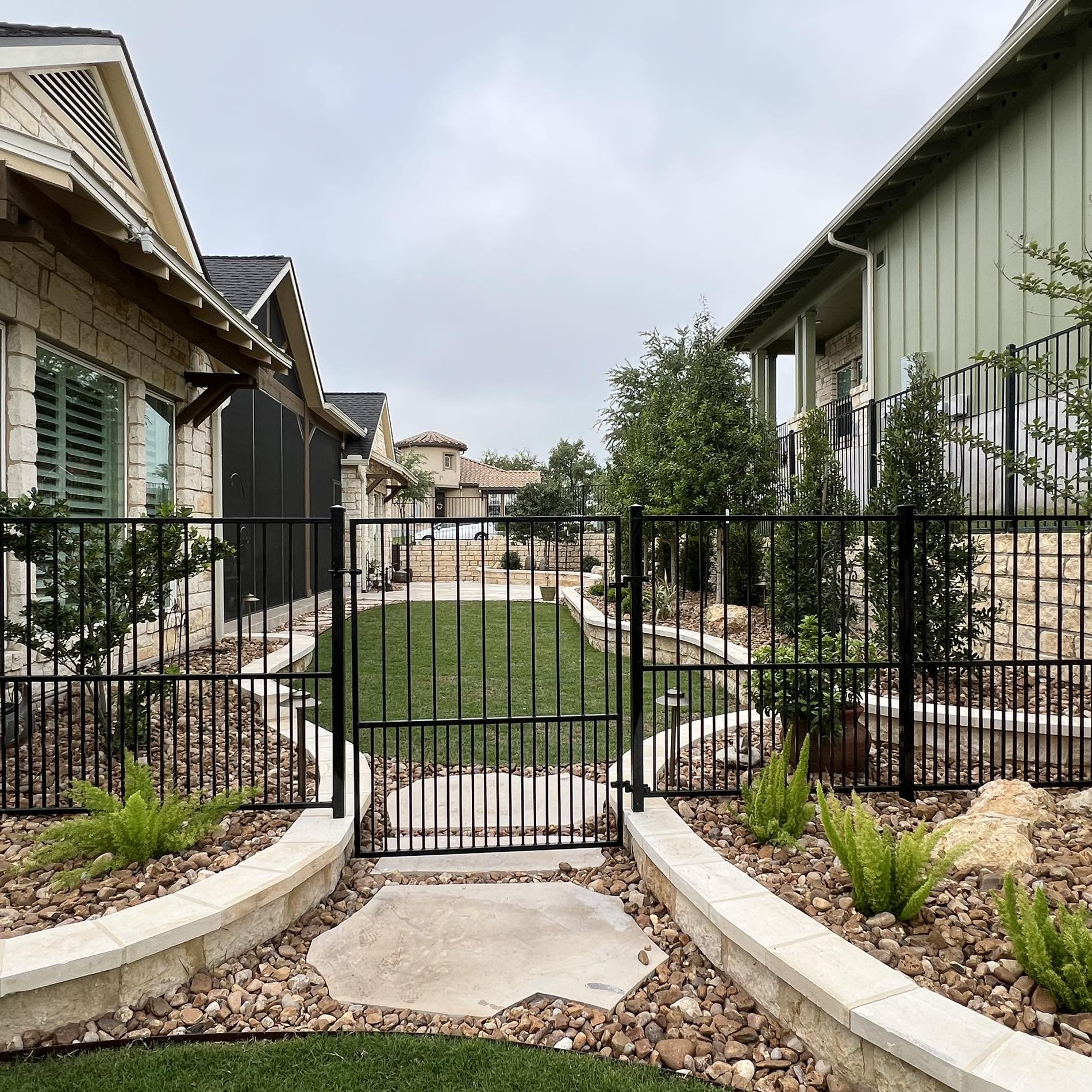 View of a backyard with a black metal fence, small gate, and landscaped garden with rocks, green plants, and a grassy lawn area, surrounded by houses with stone and siding exteriors.
