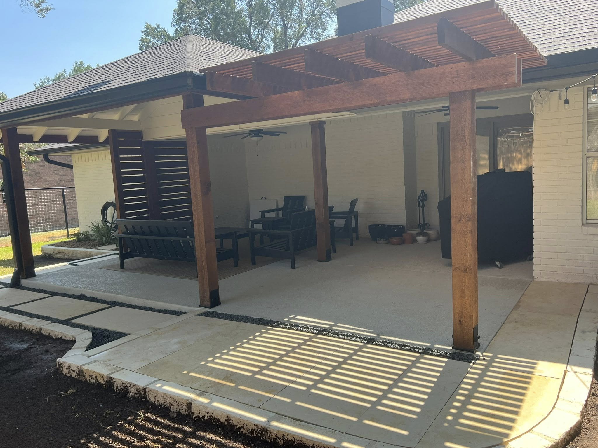 A covered outdoor patio with seating furniture, wooden support beams, a ceiling fan, hanging string lights, and a barbecue grill, attached to a house with white brick walls and a window.