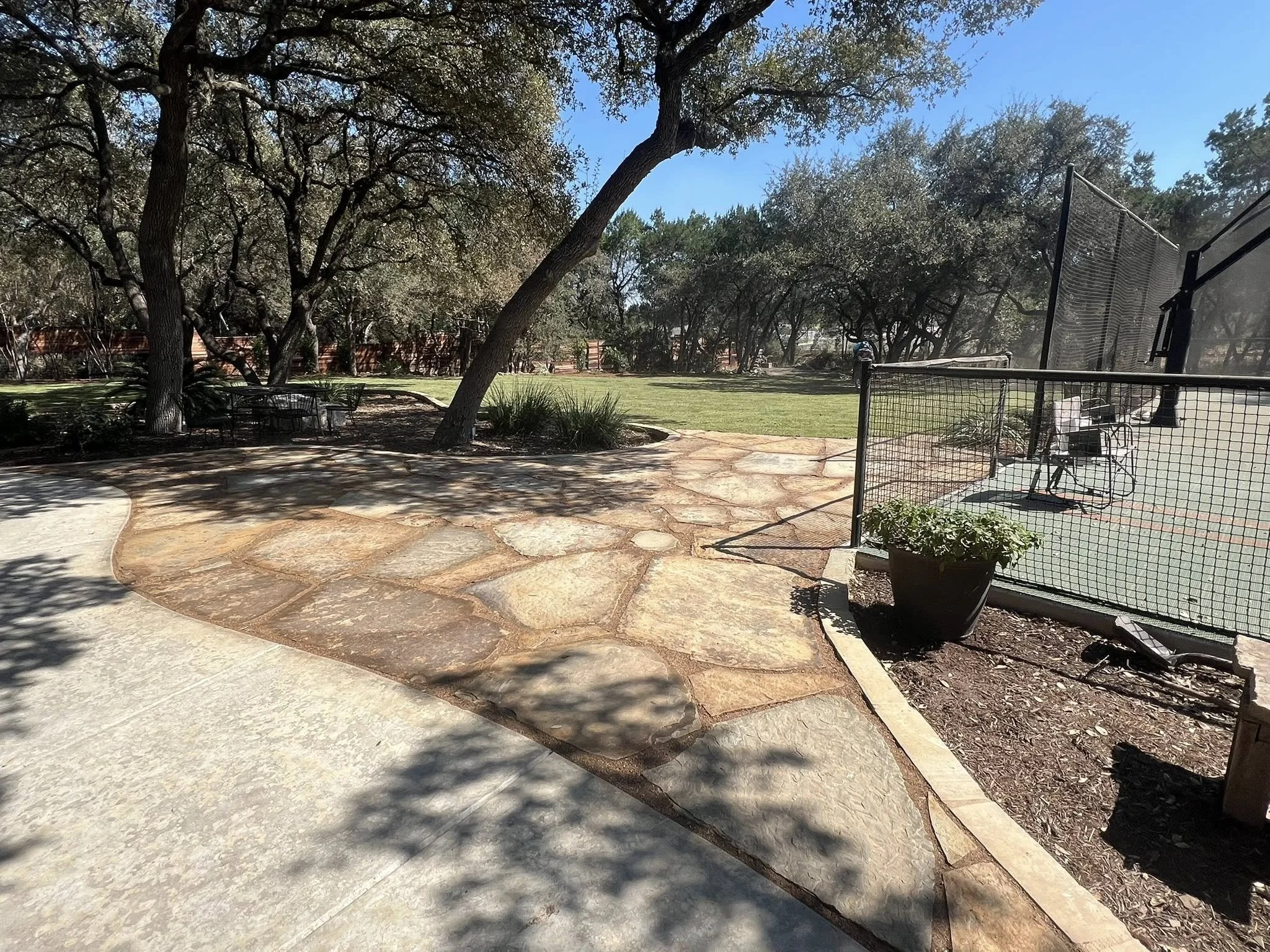 A sunny outdoor park with a stone pathway, green grass, several trees, a fenced tennis court with sports equipment, and a clear blue sky.