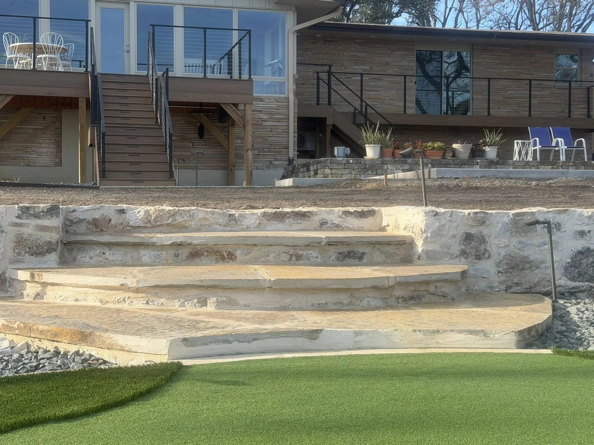 A backyard scene with stone steps leading up to a deck with chairs, potted plants, and large glass windows on a modern house. The foreground features a green artificial lawn.