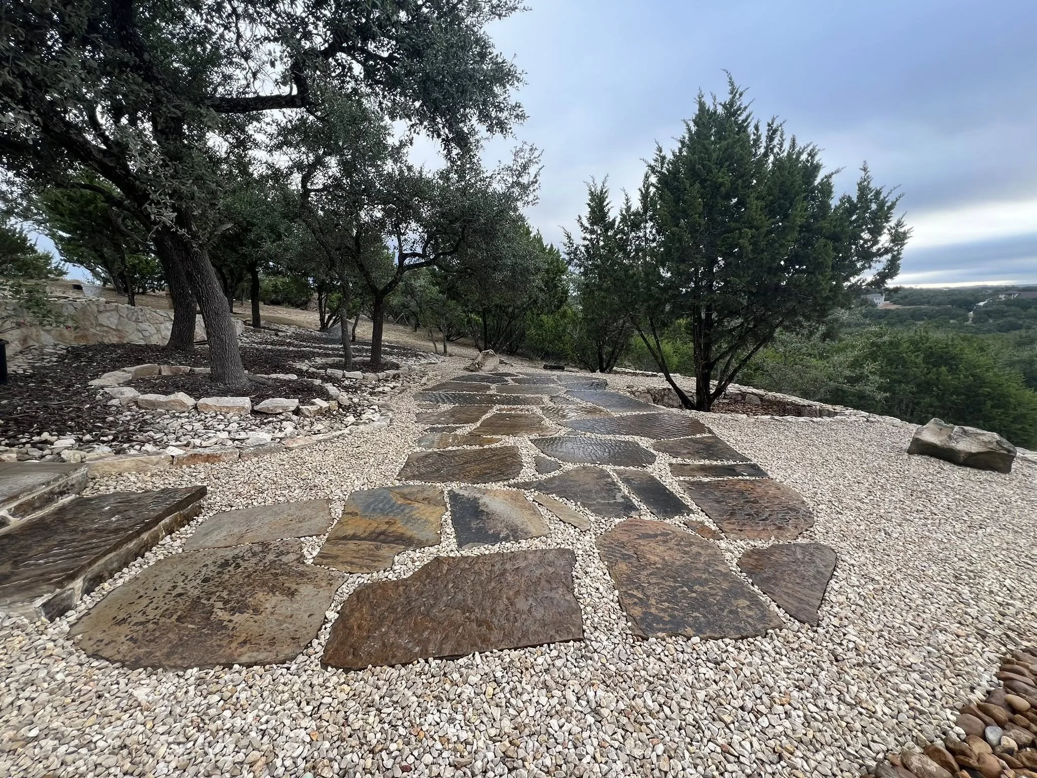 Stone pathway winding through a landscaped area with trees and gravel ground cover, overlooking a distant forested landscape under a cloudy sky.