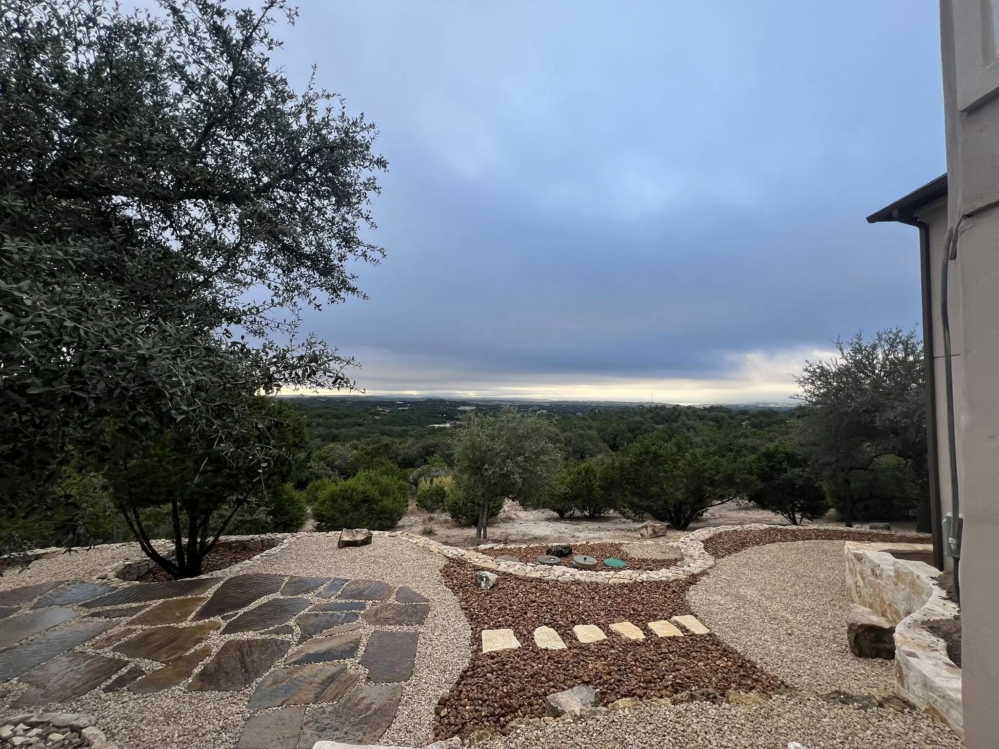 A landscape view with a cloudy sky over a desert or semi-arid area. In the foreground, there is a gravel and stone pathway, with some trees and bushes, and part of a building is visible on the right side.