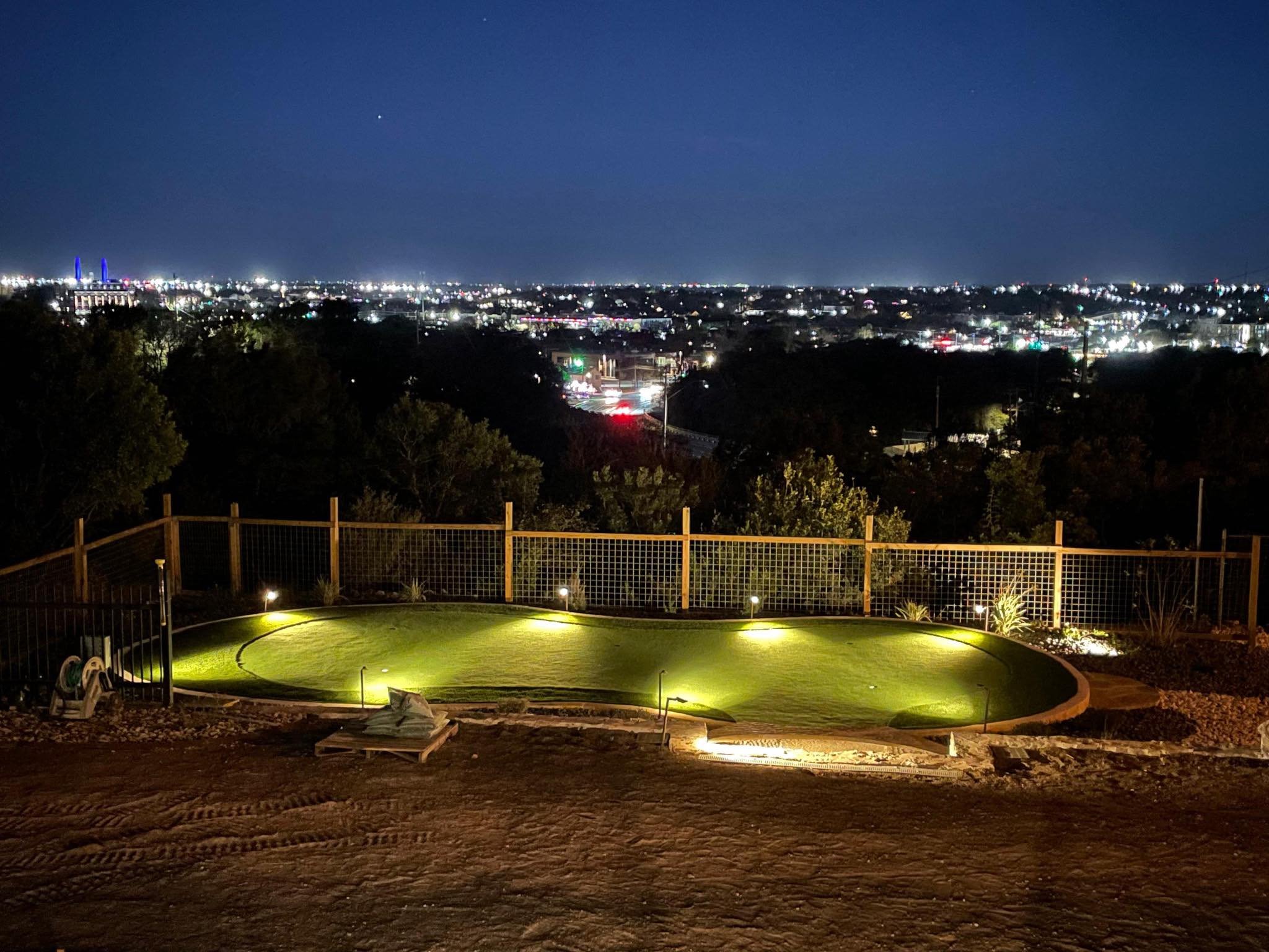 Nighttime view of a backyard with a small, circular golf putting green illuminated by ground lights, overlooking a city skyline filled with bright lights under a dark sky.
