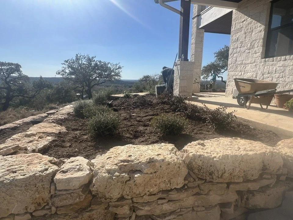 Person working on gardening or landscaping near a stone wall outside a house with an off-white brick exterior. There is a wheelbarrow and a clay pot nearby, with trees and a clear blue sky in the background.