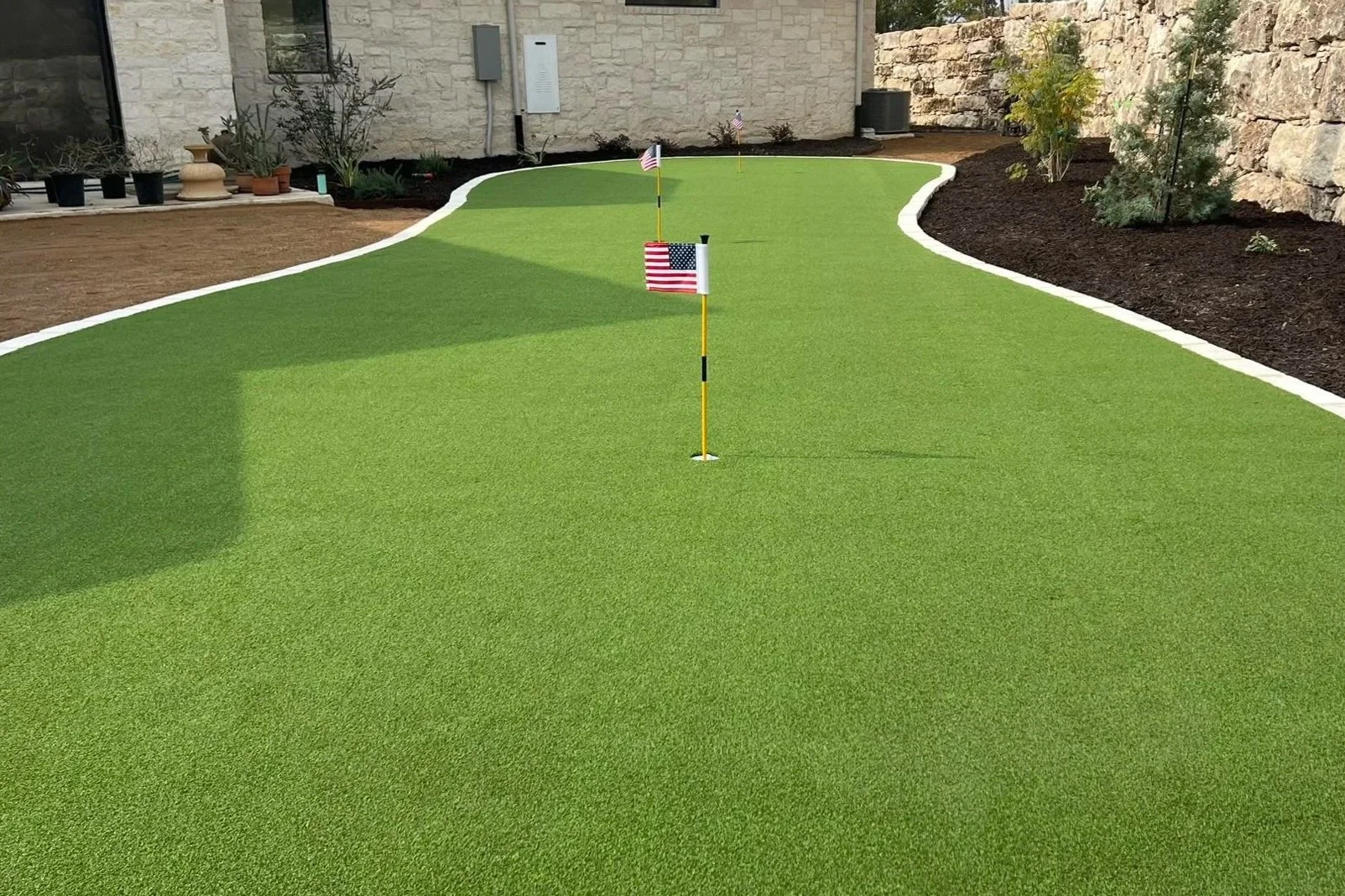 Mini golf putting green with three small American flags on yellow poles, surrounded by a stone wall and garden plants.