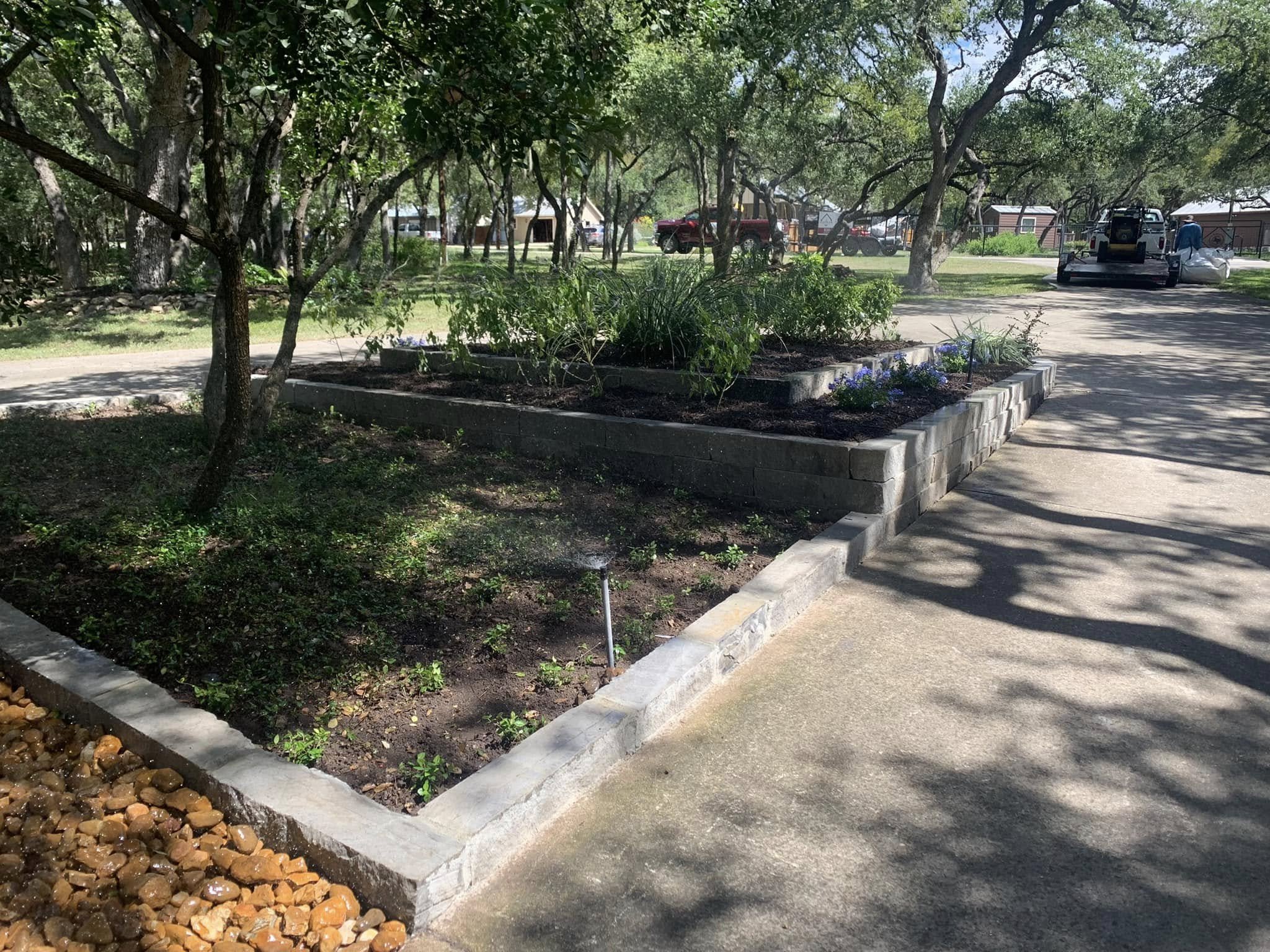 A small garden bed with plants and flowers, bordered by concrete blocks, located beside a sidewalk in a park or residential area, with trees casting shadows on the ground.