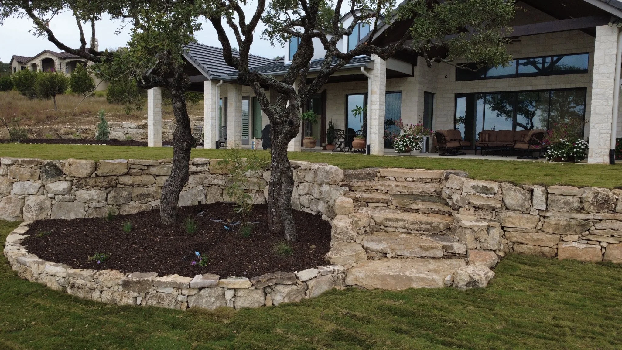 A backyard with a raised stone garden bed containing a tree and small plants, with stone steps leading up to a grassy patio area in front of a house with large windows and outdoor furniture.