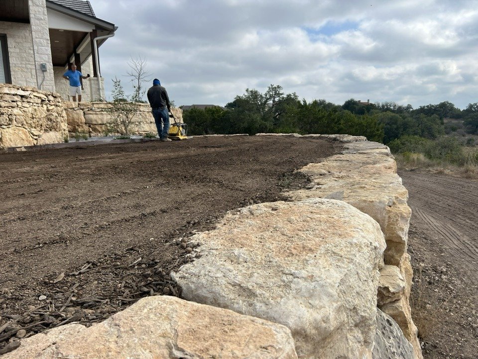 Construction workers installing a layer of soil and gravel on a raised stone patio or wall in a backyard with a house and trees in the background.