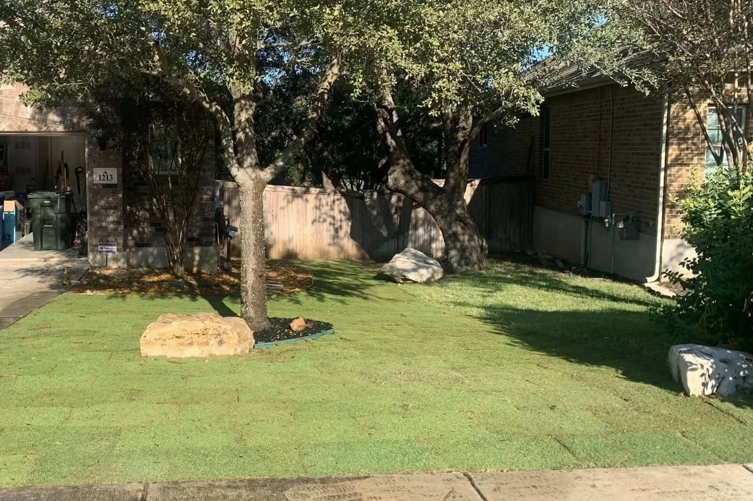 Residential backyard with trimmed grass, trees, rocks, and a brick house with visible power meters and pipes.
