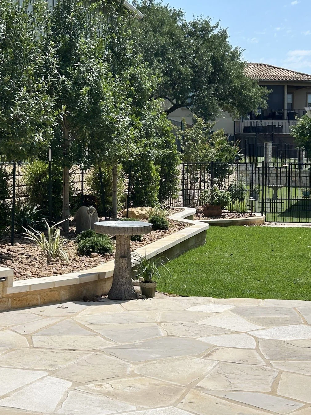 A backyard with a stone patio, a birdbath, potted plants, a green lawn, a landscaped garden bed with shrubs and trees, and a black metal fence.