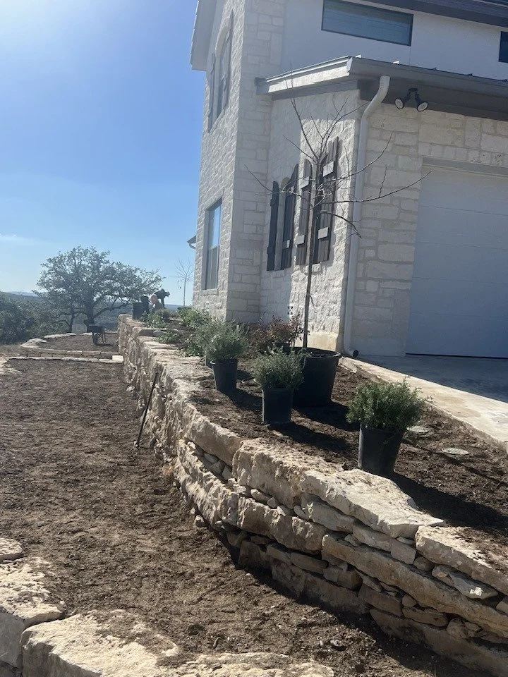 Part of a modern house with a white stone exterior, windows with black shutters, and a garage door. In front, there is a stone retaining wall with potted plants on top, and a dirt pathway running alongside.